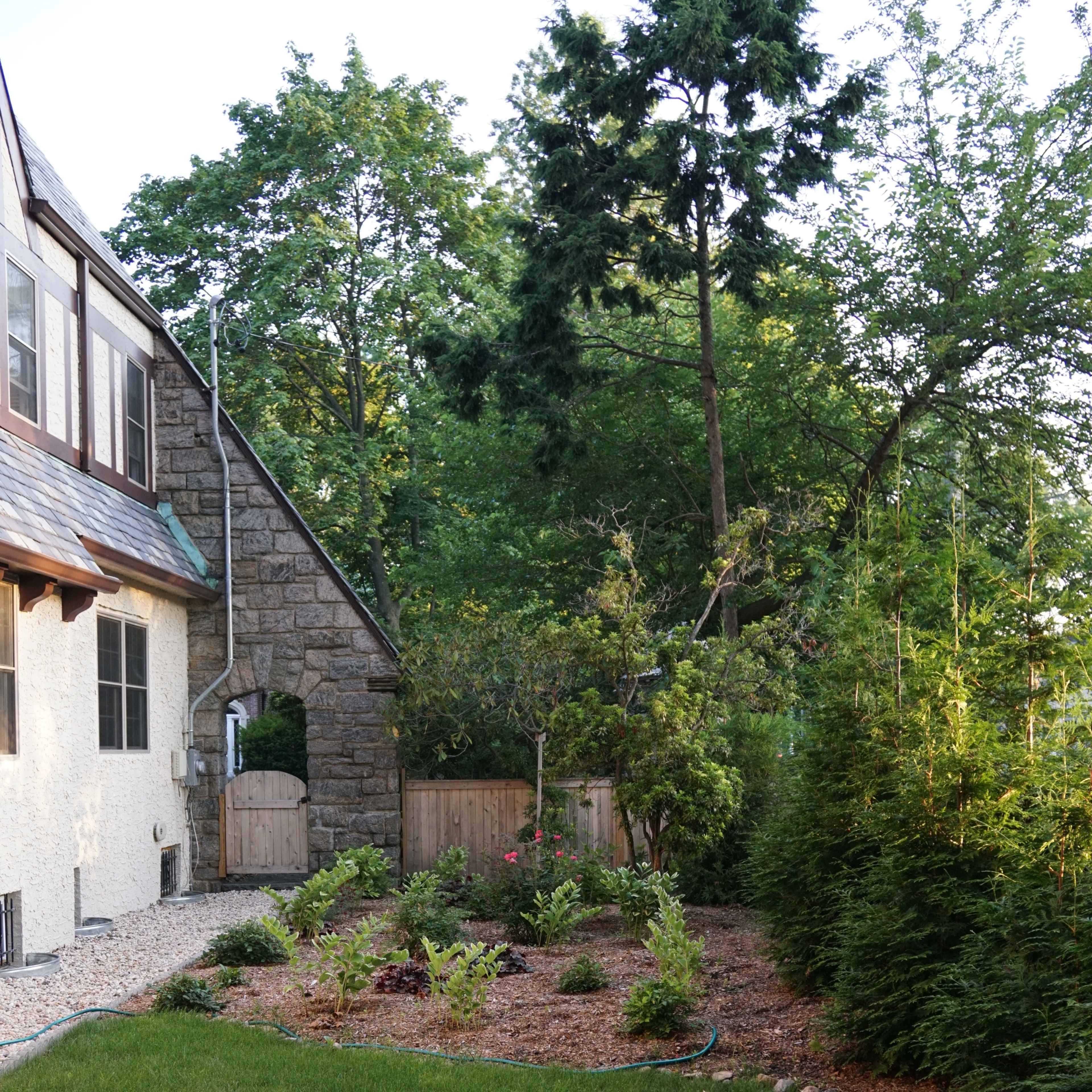 A well-maintained garden with grass and small shrubs is located beside a stone house, flanked by trees and a wooden fence.