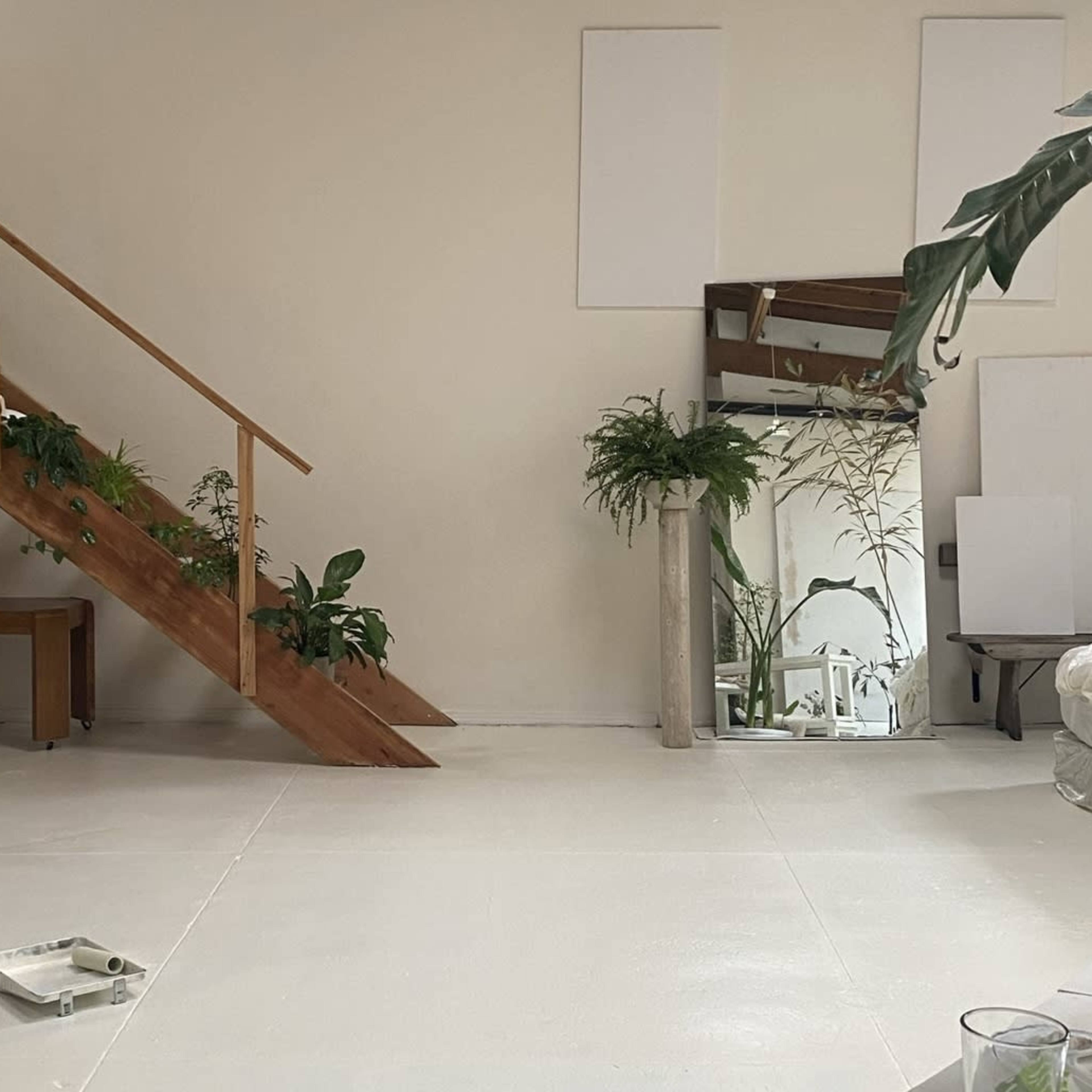 A minimalist interior space featuring a wooden staircase with plants, a small wooden table, a large mirror, and several potted plants.