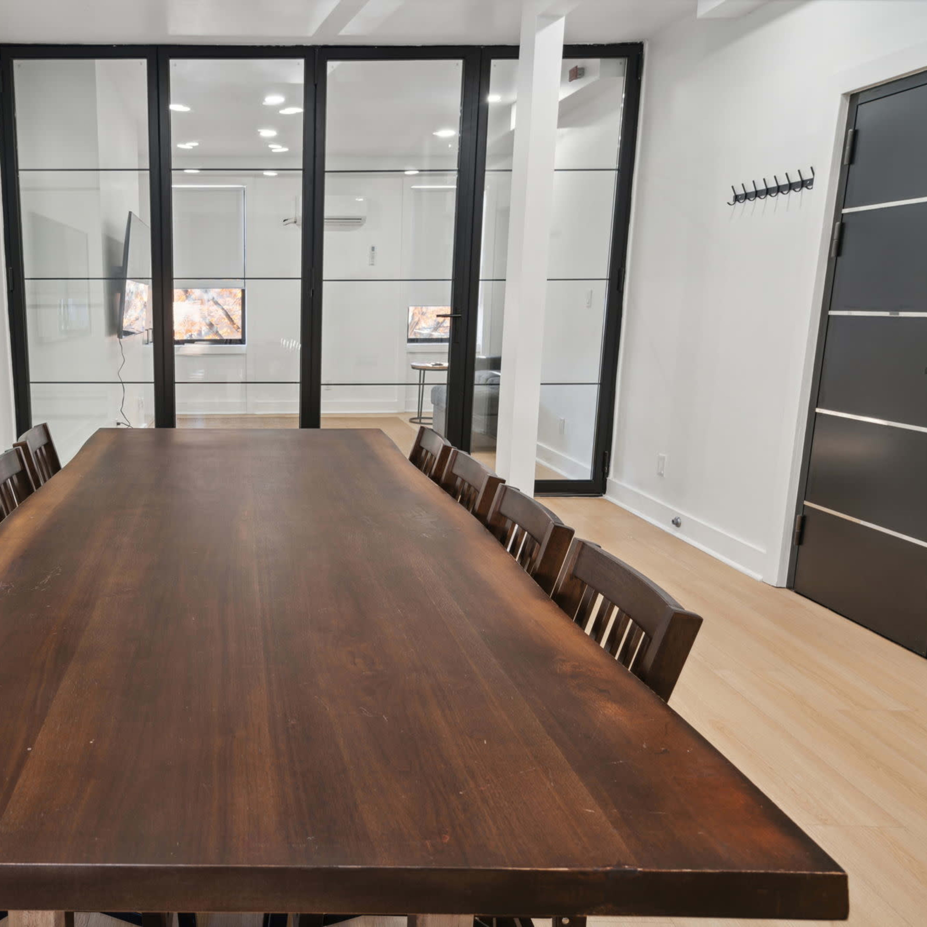 A long wooden table with eight chairs is positioned in a well-lit room separated by glass walls, featuring two screens visible in the background.