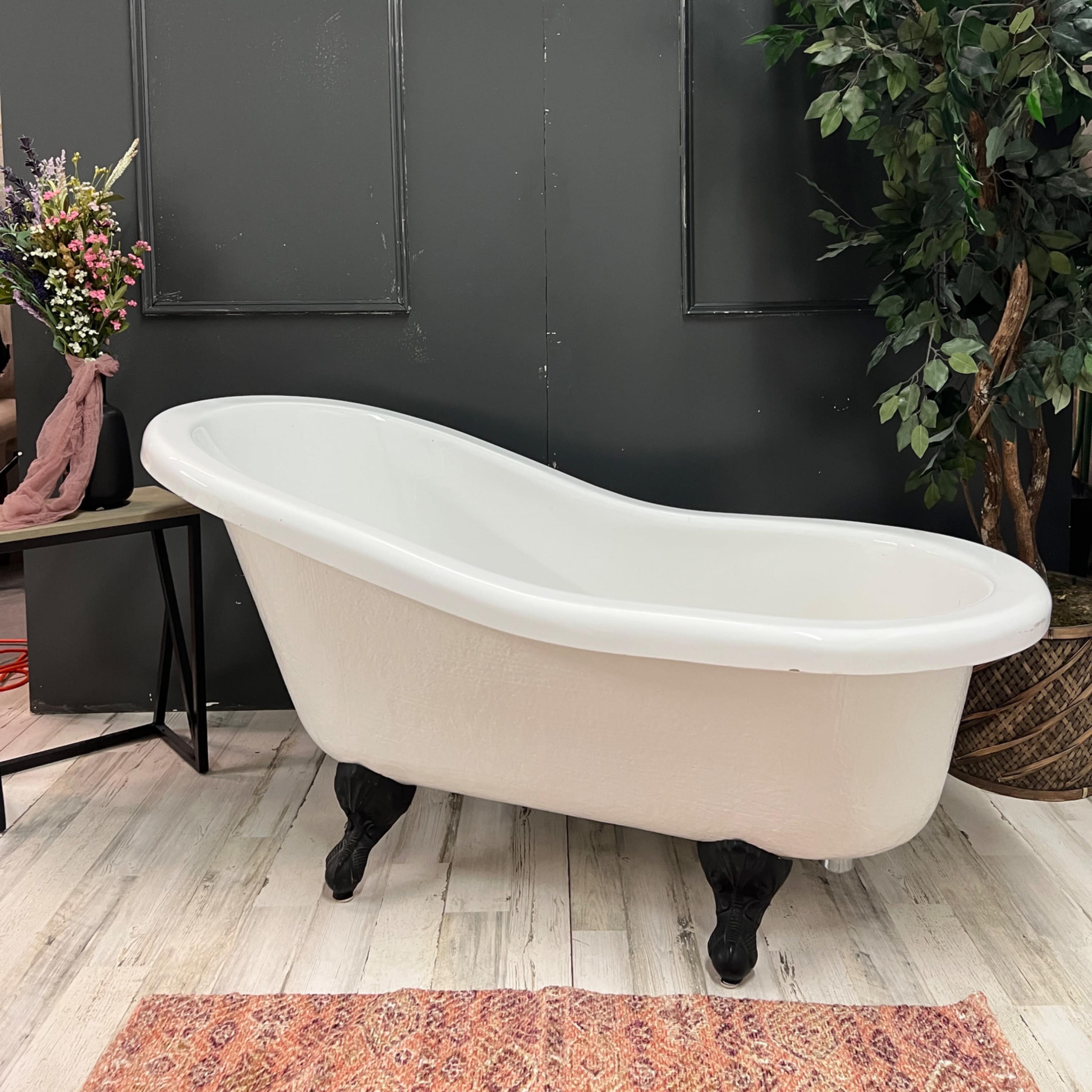 A white vintage-style bathtub on claw feet positioned against a dark wall, accompanied by a potted plant and a floral arrangement on a small table.