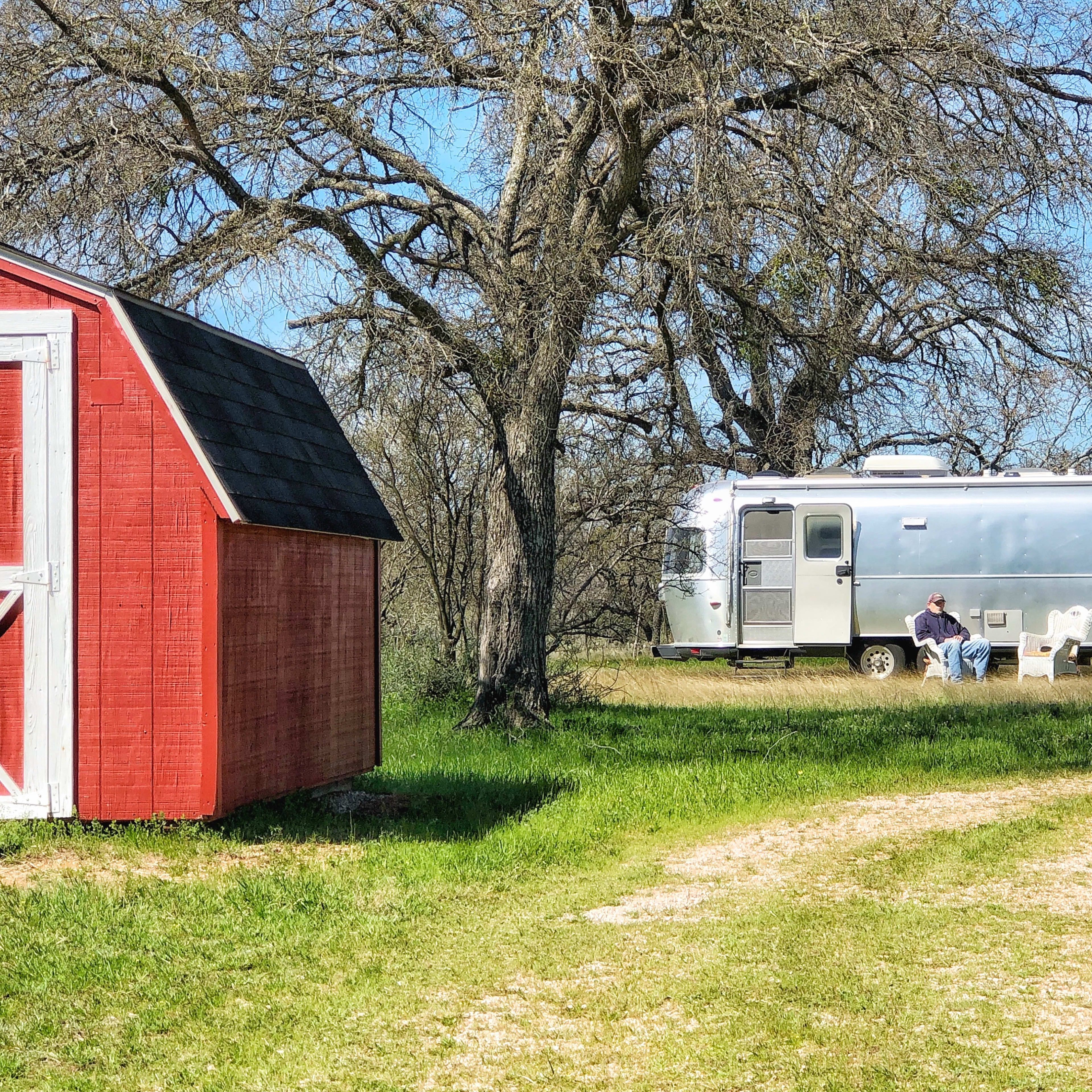 A silver Airstream trailer is parked under trees, beside a red shed, on a grassy path.