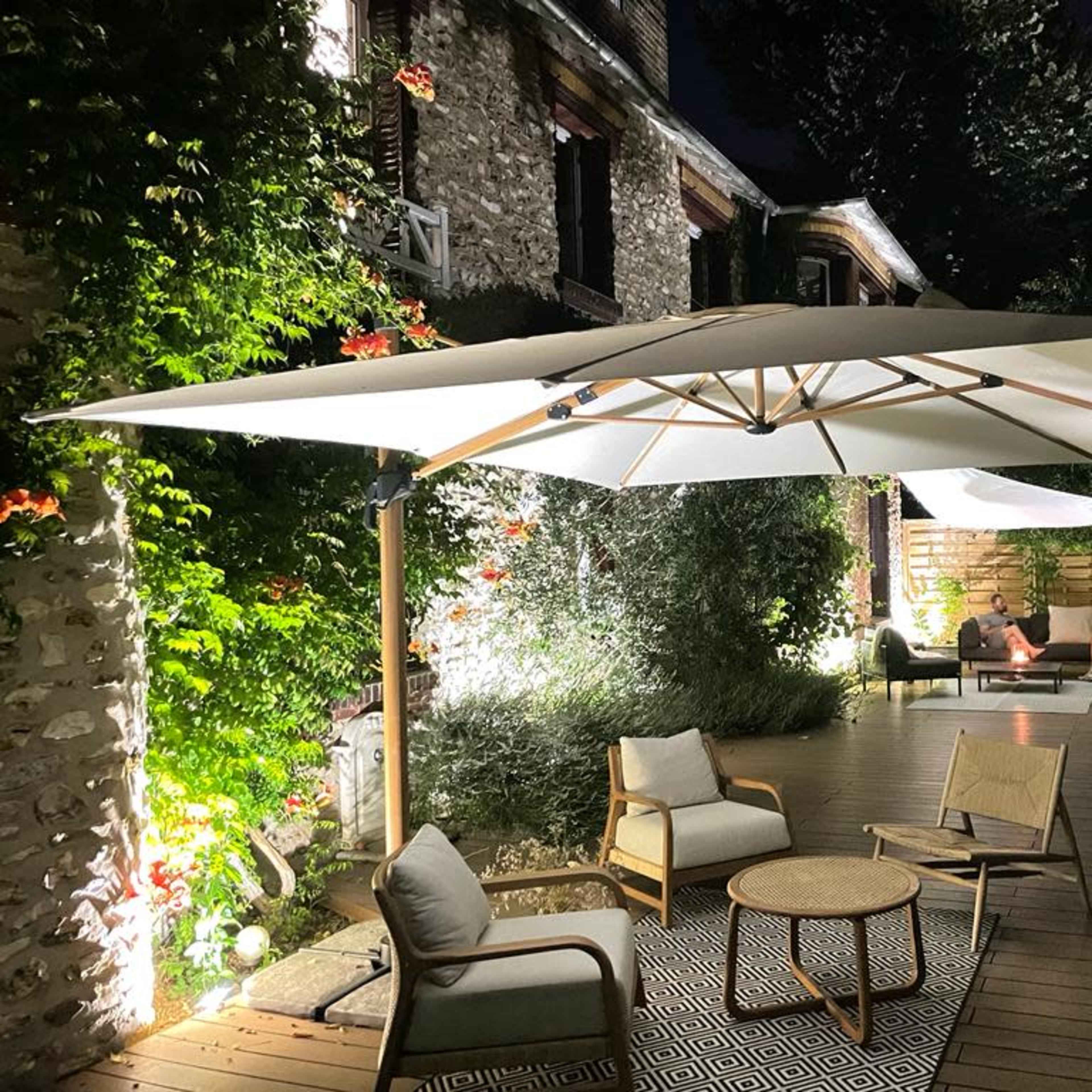 A patio area is illuminated at night, featuring a large umbrella over a wooden deck with seating and plants.