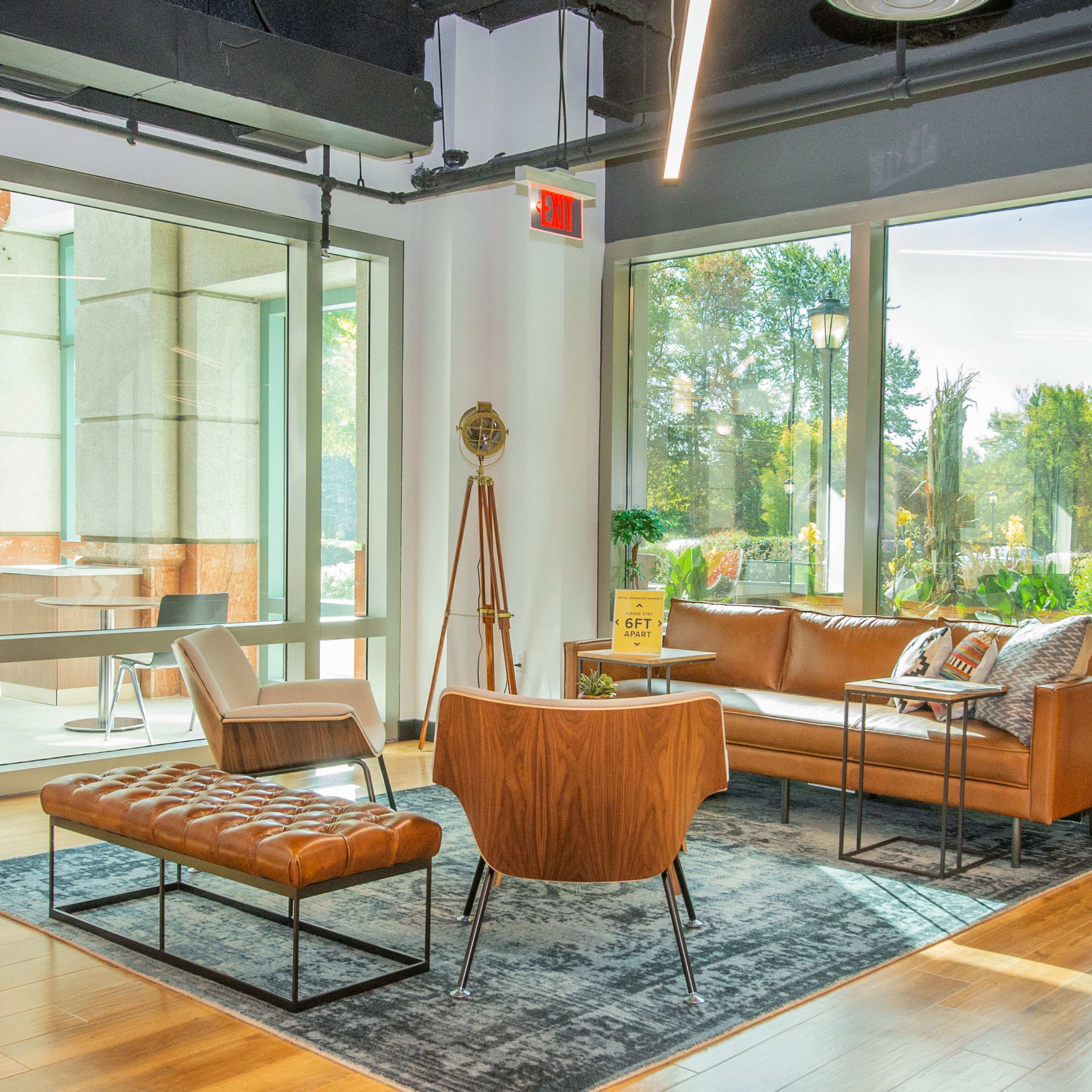 A modern lounge area with a brown leather sofa, an armchair, a footrest, and large windows overlooking a garden.