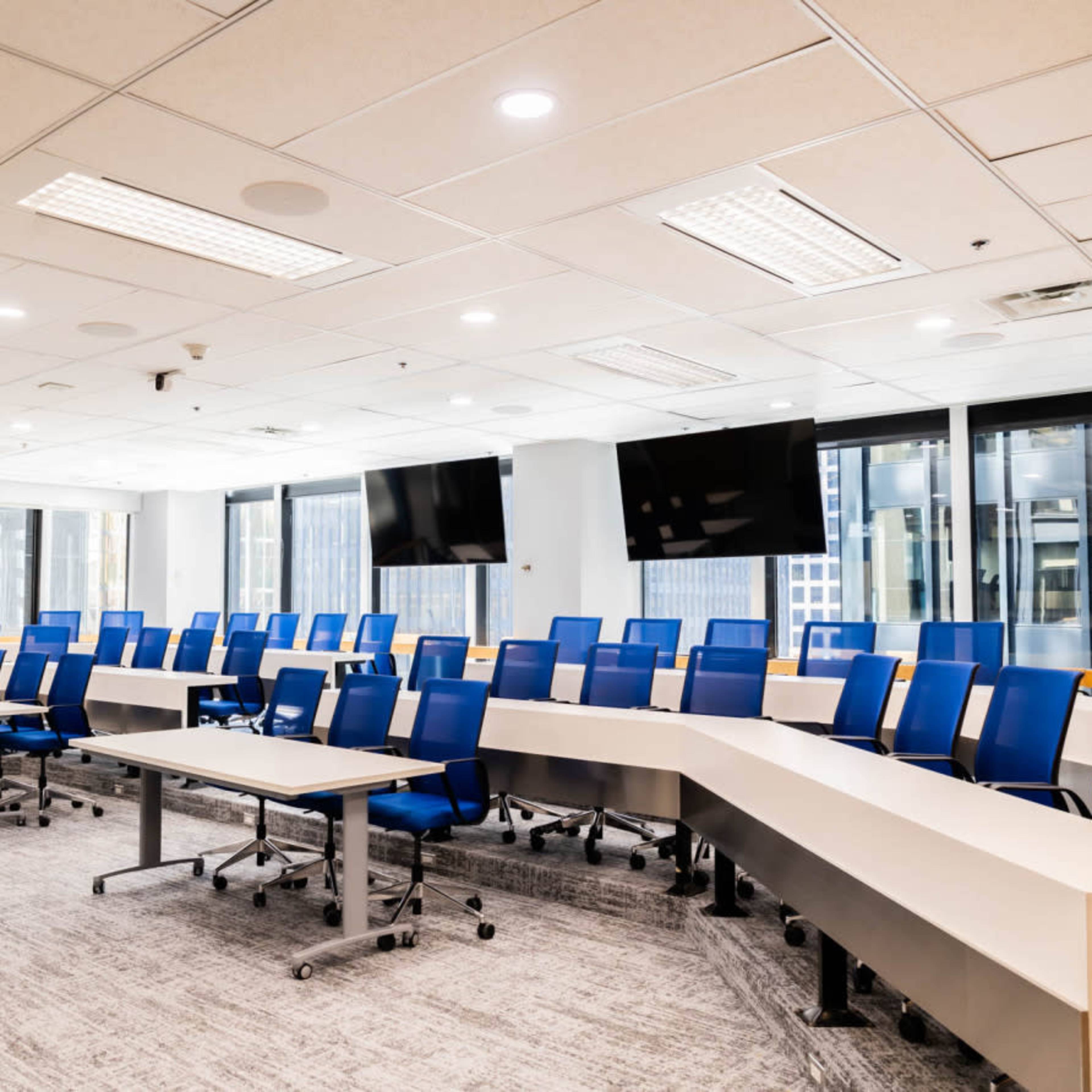 A modern conference room features multiple rows of blue chairs and tables arranged in a curved layout, with large windows and two mounted screens.