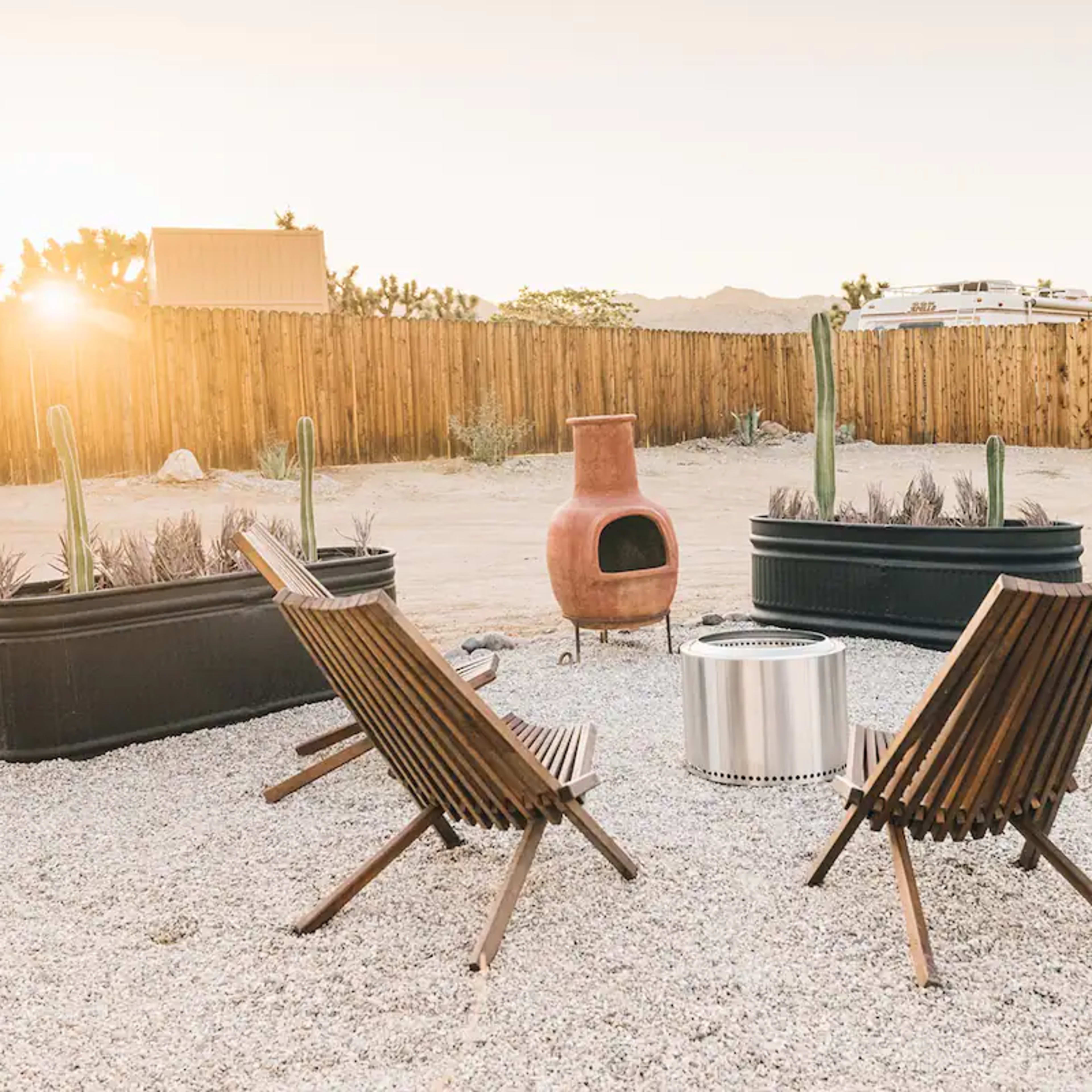 A circular seating area with four wooden chairs surrounds a fire pit in a gravel setting, framed by a wooden fence and desert plants at sunset.