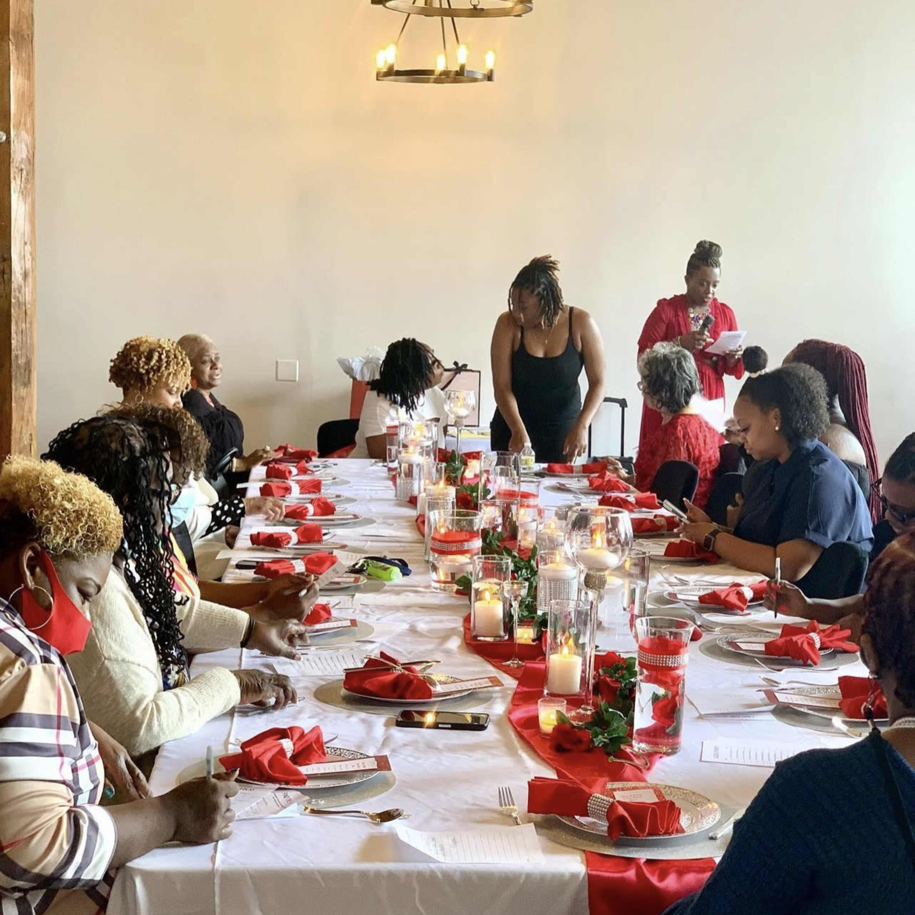 A group of women gather around a long dining table set with red napkins and candles for a meal or event.