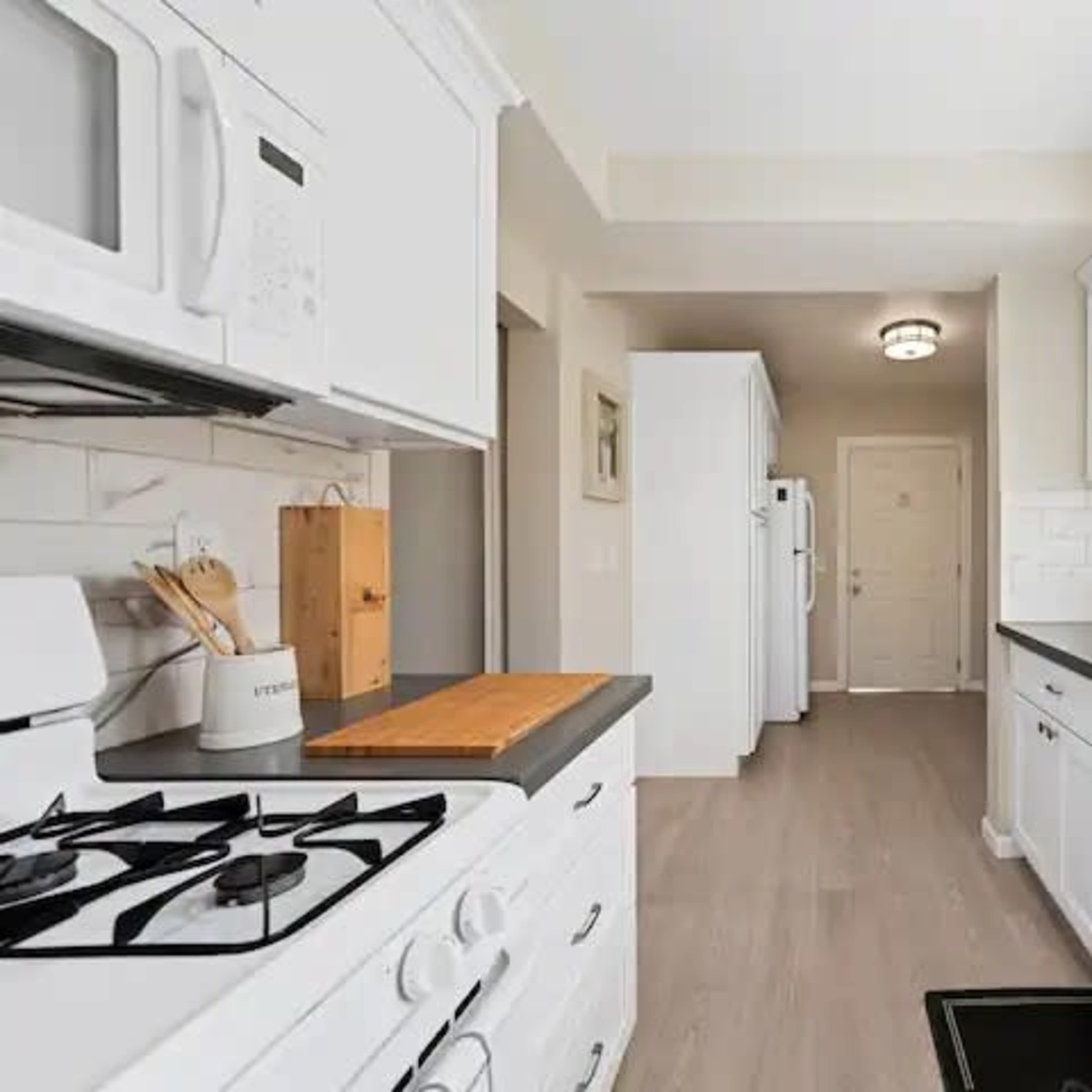 A clean, modern kitchen with white cabinetry, a gas stove, and a hallway leading to a door at the end.