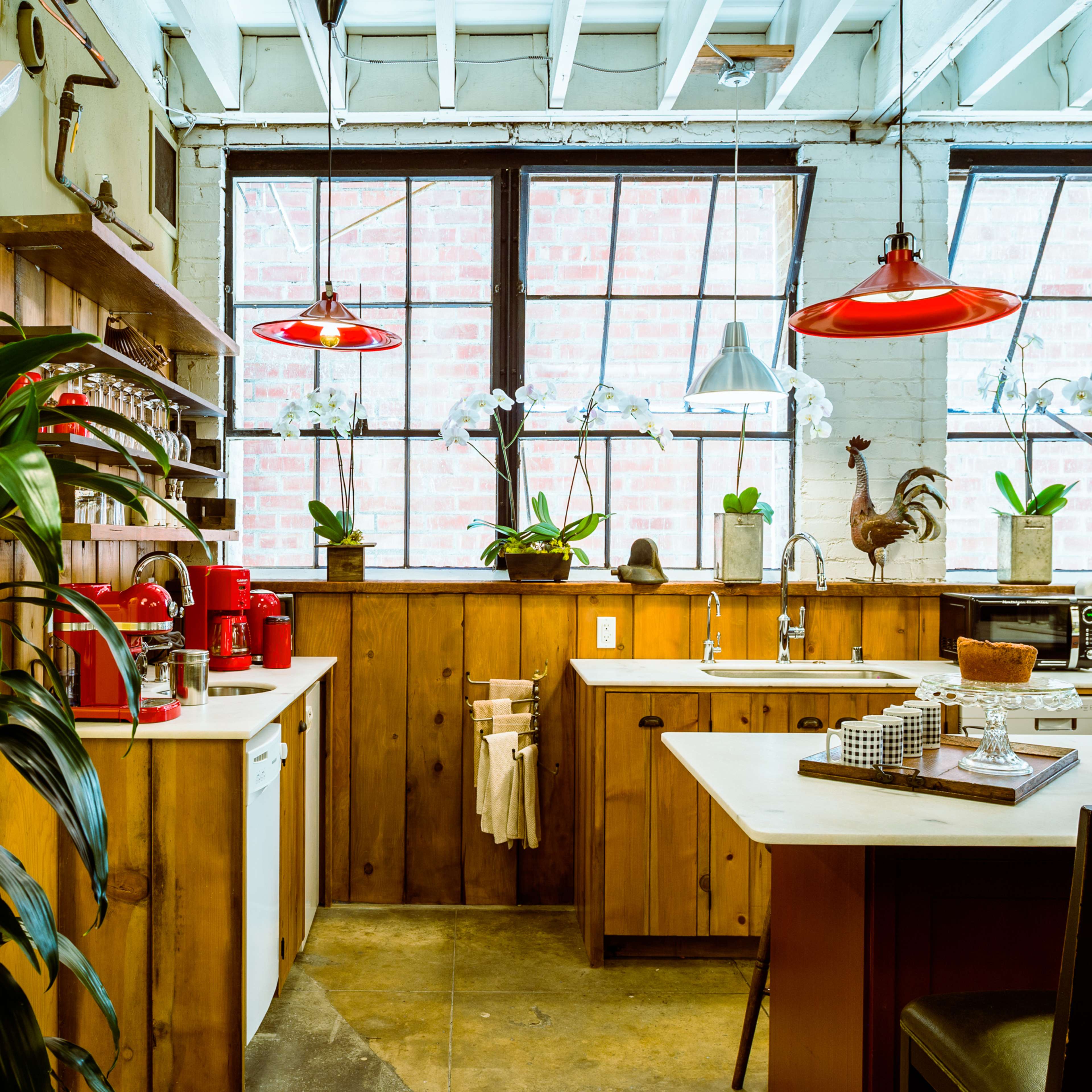 The kitchen features wooden cabinetry, a central island with seating, red pendant lights, and large windows bringing in natural light.