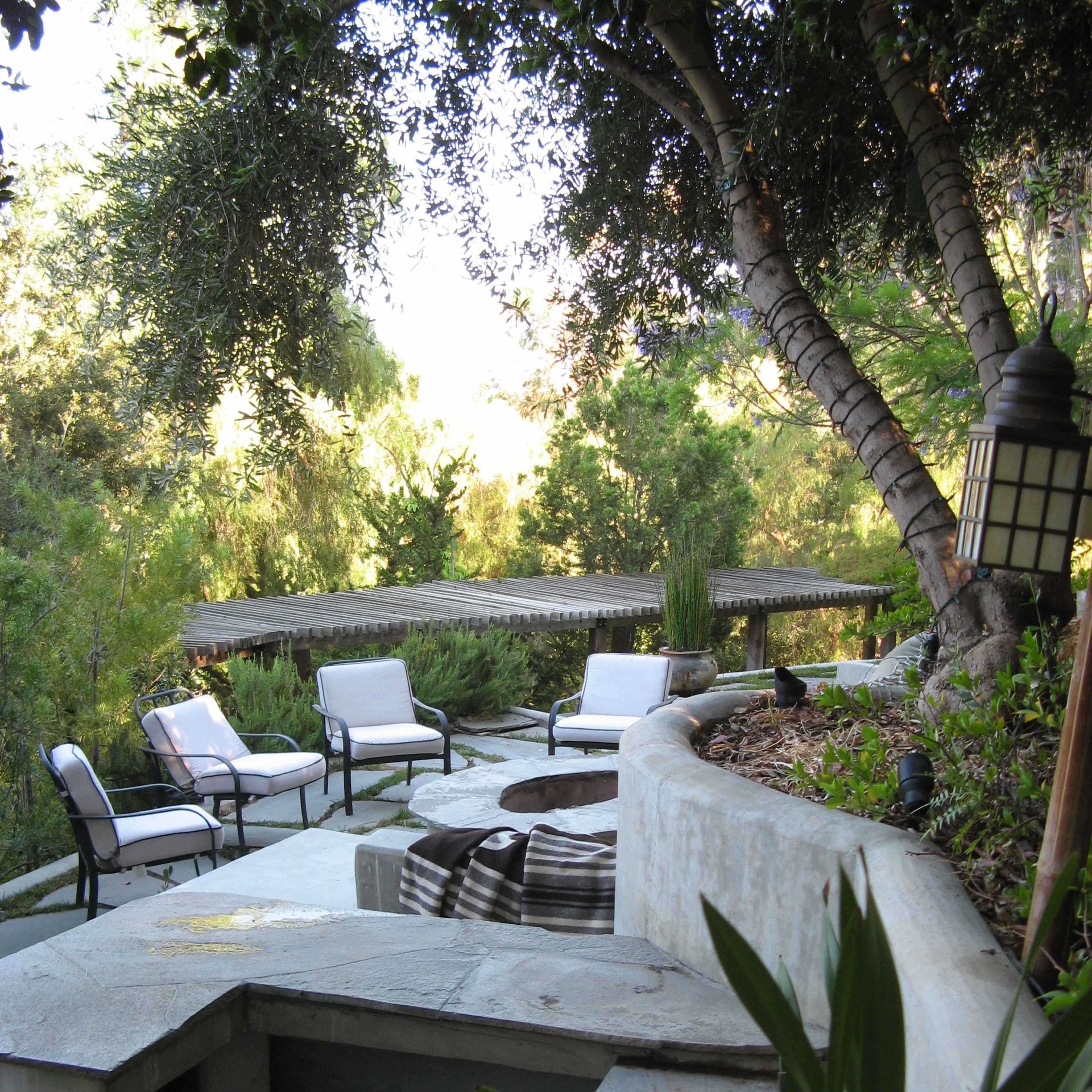 A landscaped outdoor seating area featuring several chairs arranged around a stone fire pit, with greenery and trees in the background.