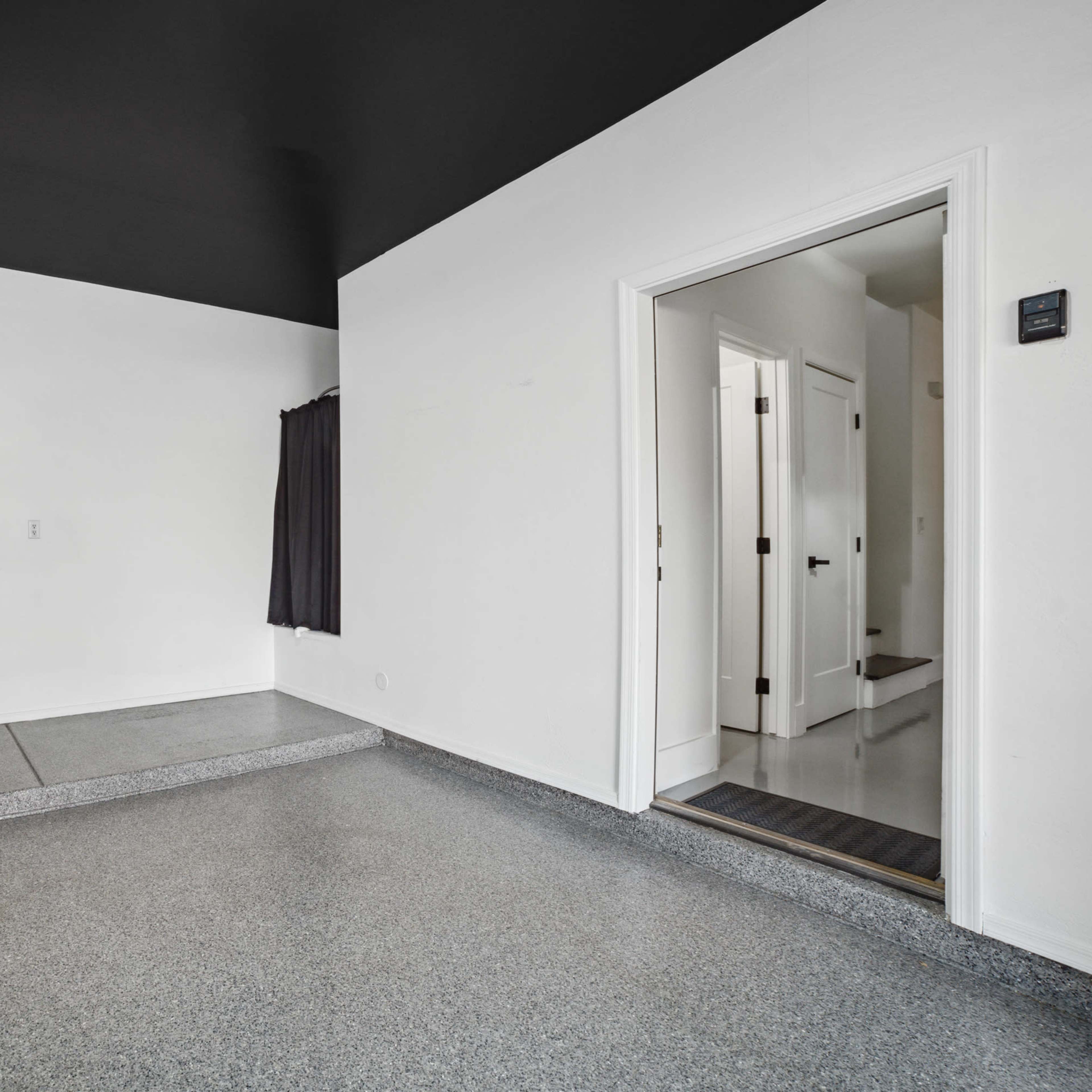 A minimalistic room with a textured gray floor, white walls, a black ceiling, a doorway leading to another room, and a wall-mounted item on the left.