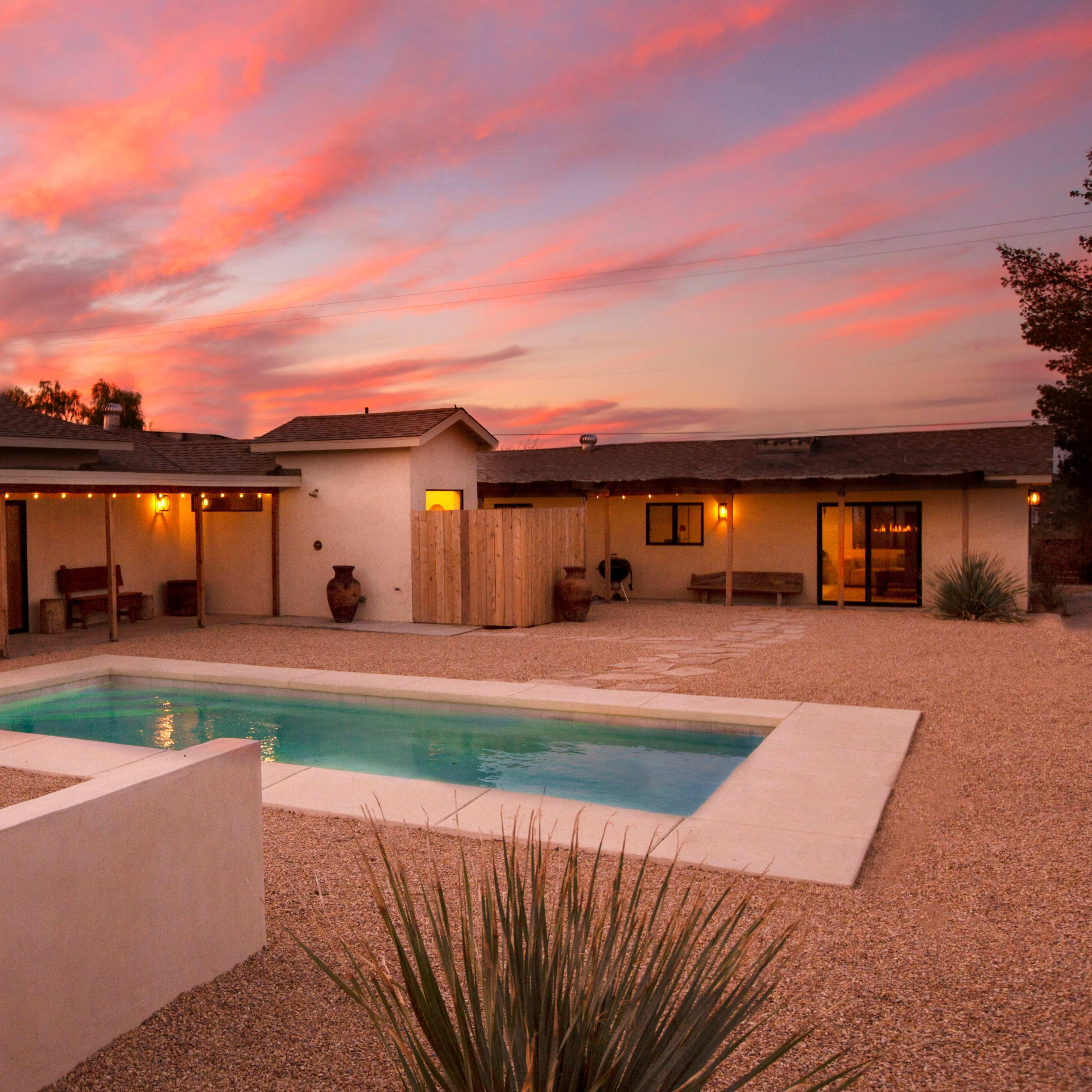 A modern home with a pool and fire feature is surrounded by a gravel courtyard under a sunset sky.