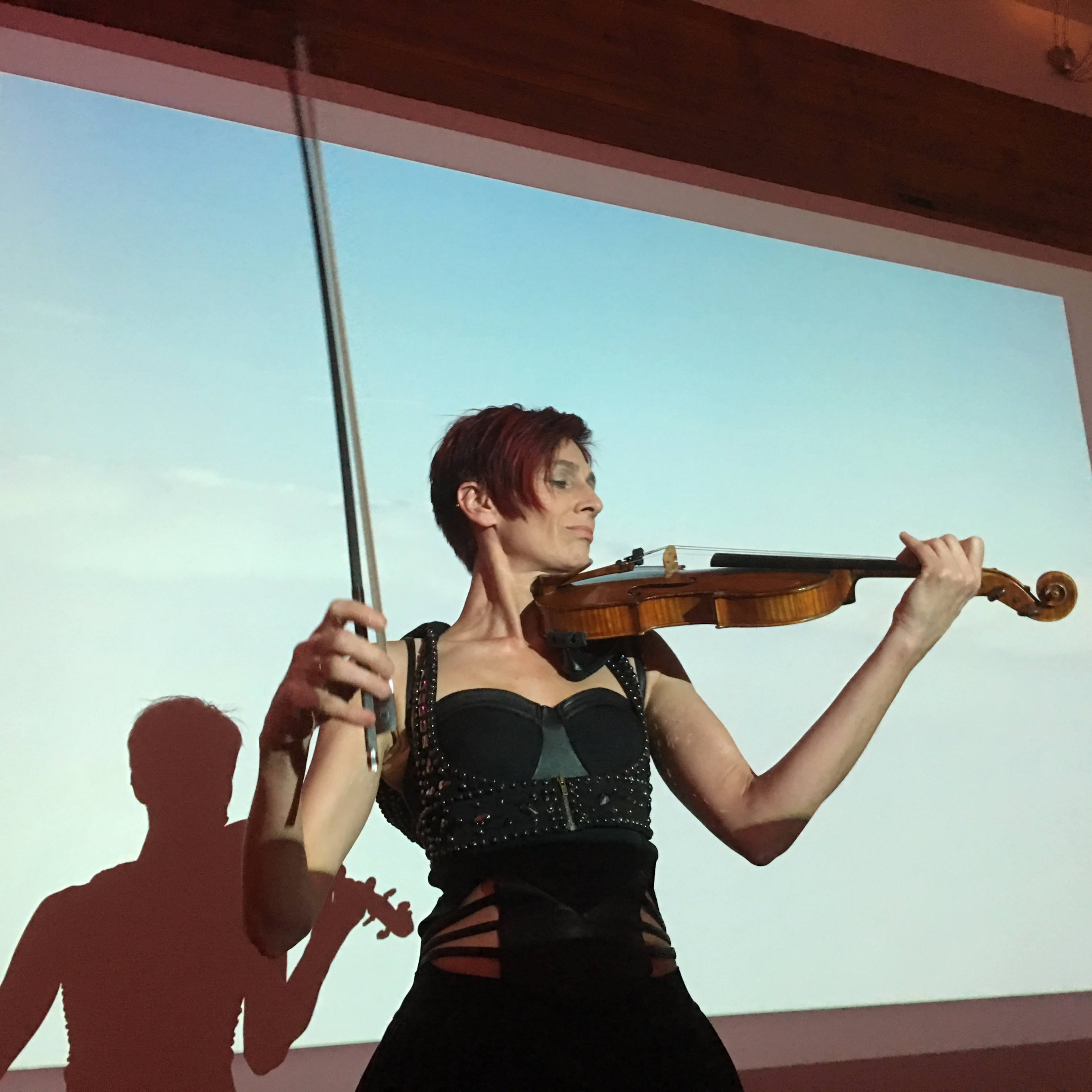A woman plays the violin while performing against a large screen displaying a clear sky.