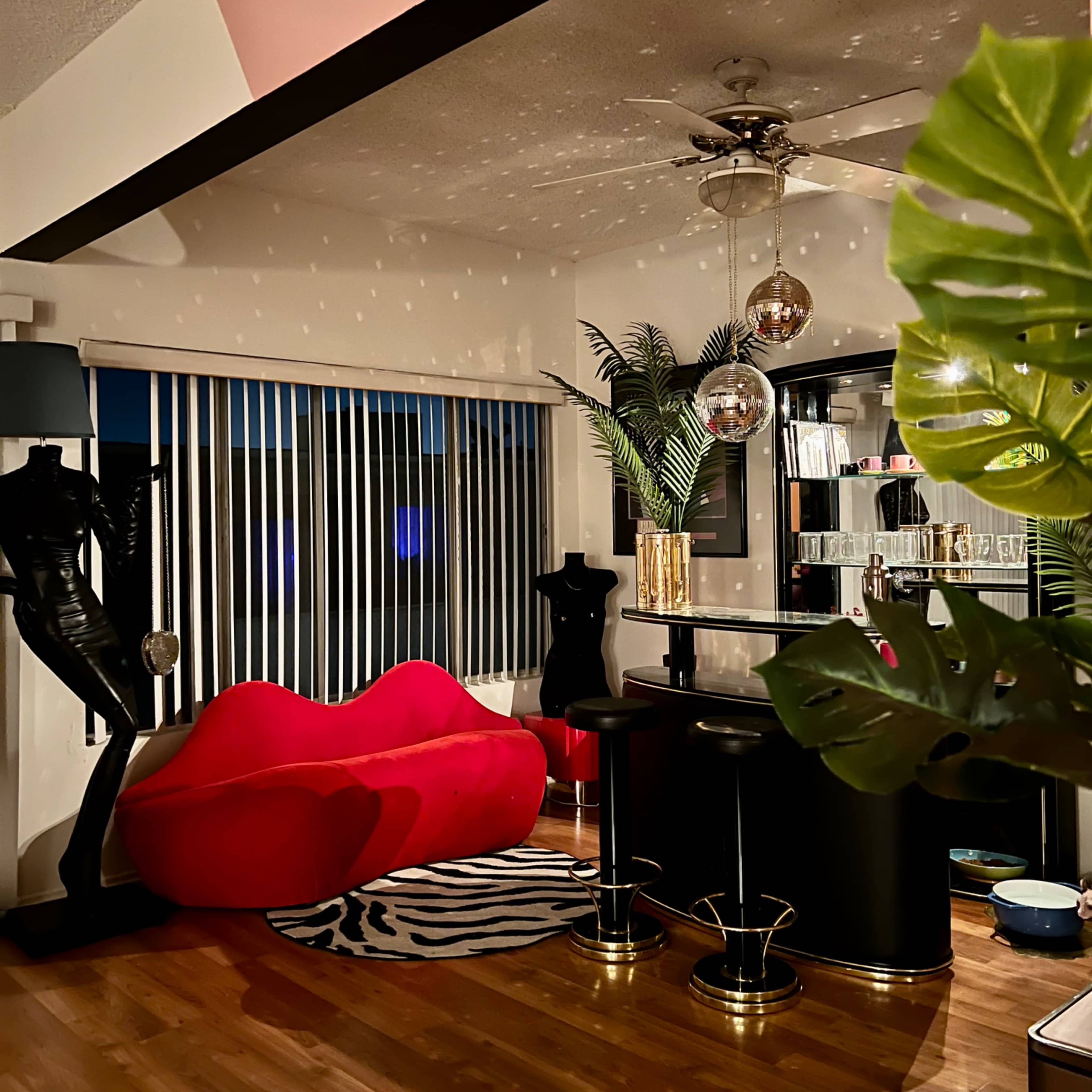 A stylish living area featuring a red couch, a black bar with stools, and decorative plants, illuminated by a ceiling fan and pendant lights.
