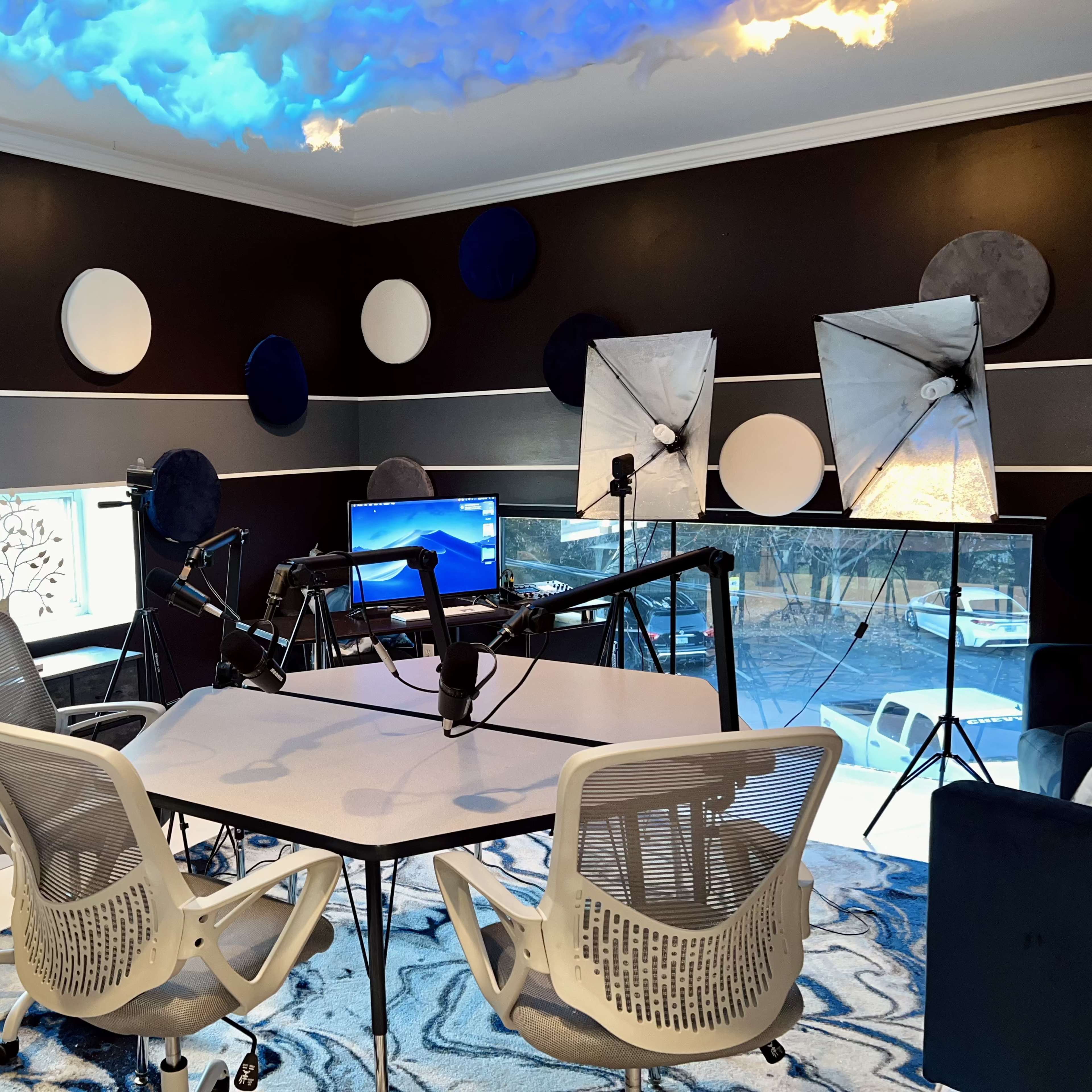 A modern recording studio with a white table, microphones, and lighting equipment, featuring a blue and black color scheme with decorative wall elements.