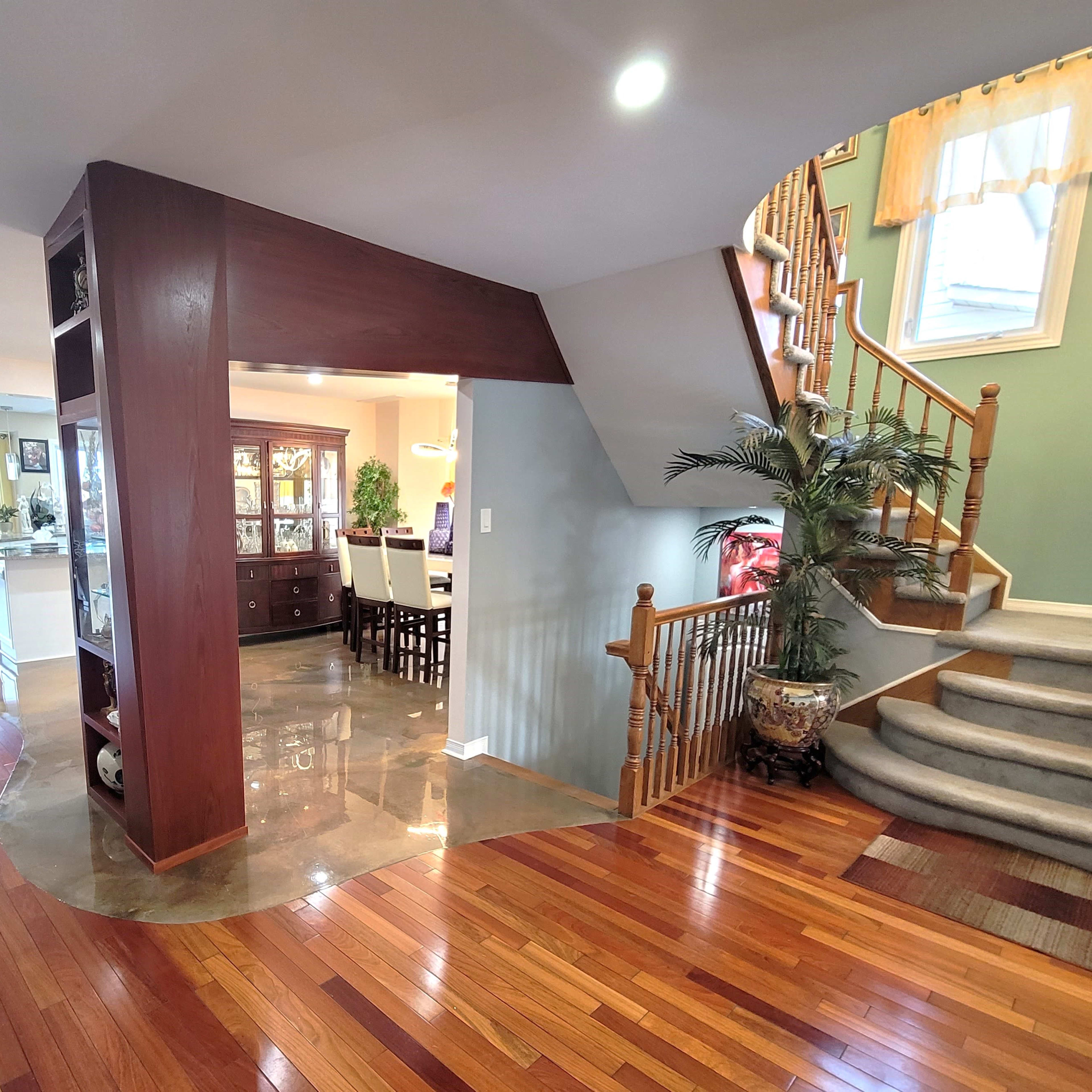A well-lit foyer with polished wooden floors, a staircase on the right, and a view of a dining area through an archway.