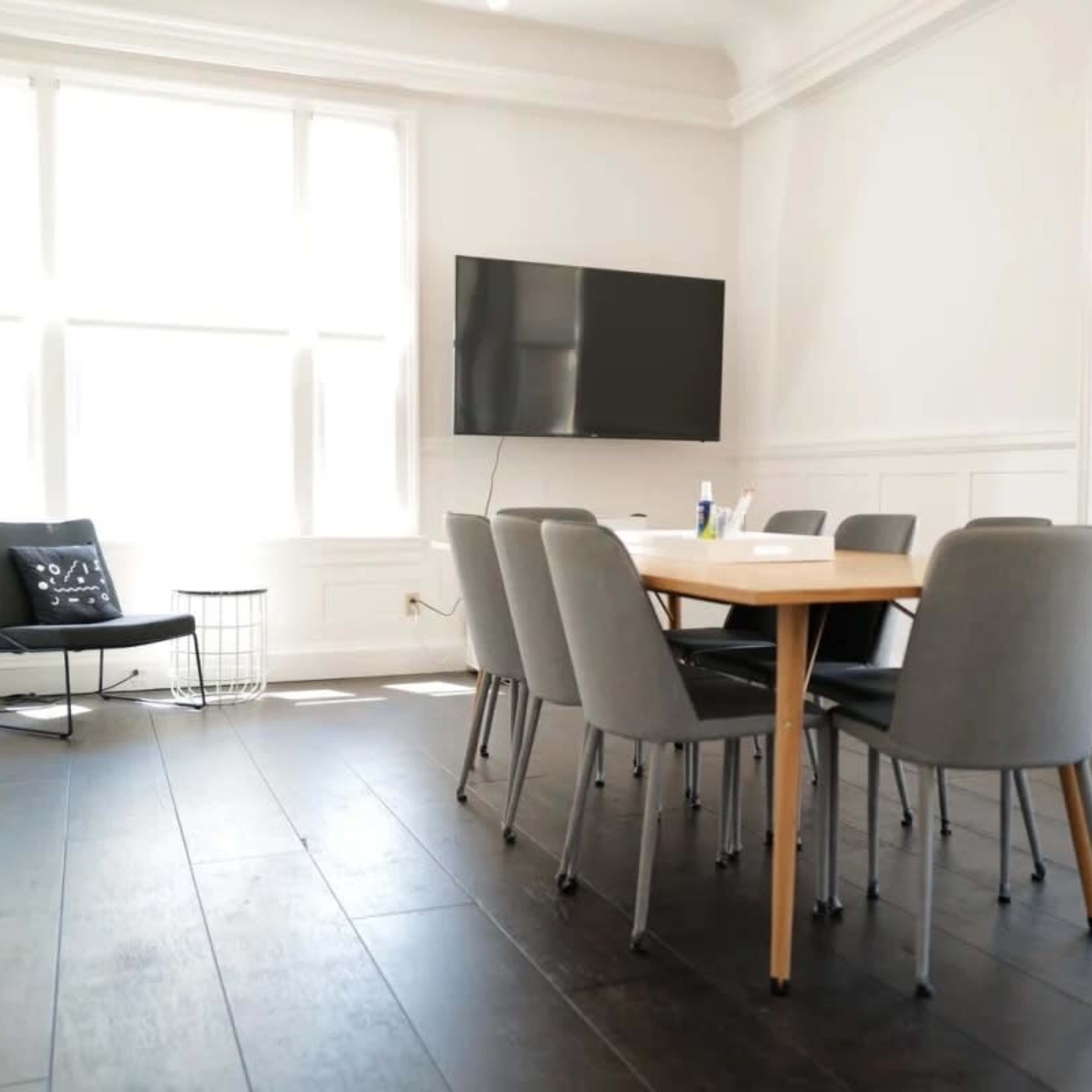 A modern meeting room features a wooden table surrounded by six gray chairs, with a television mounted on the wall and large windows allowing natural light.