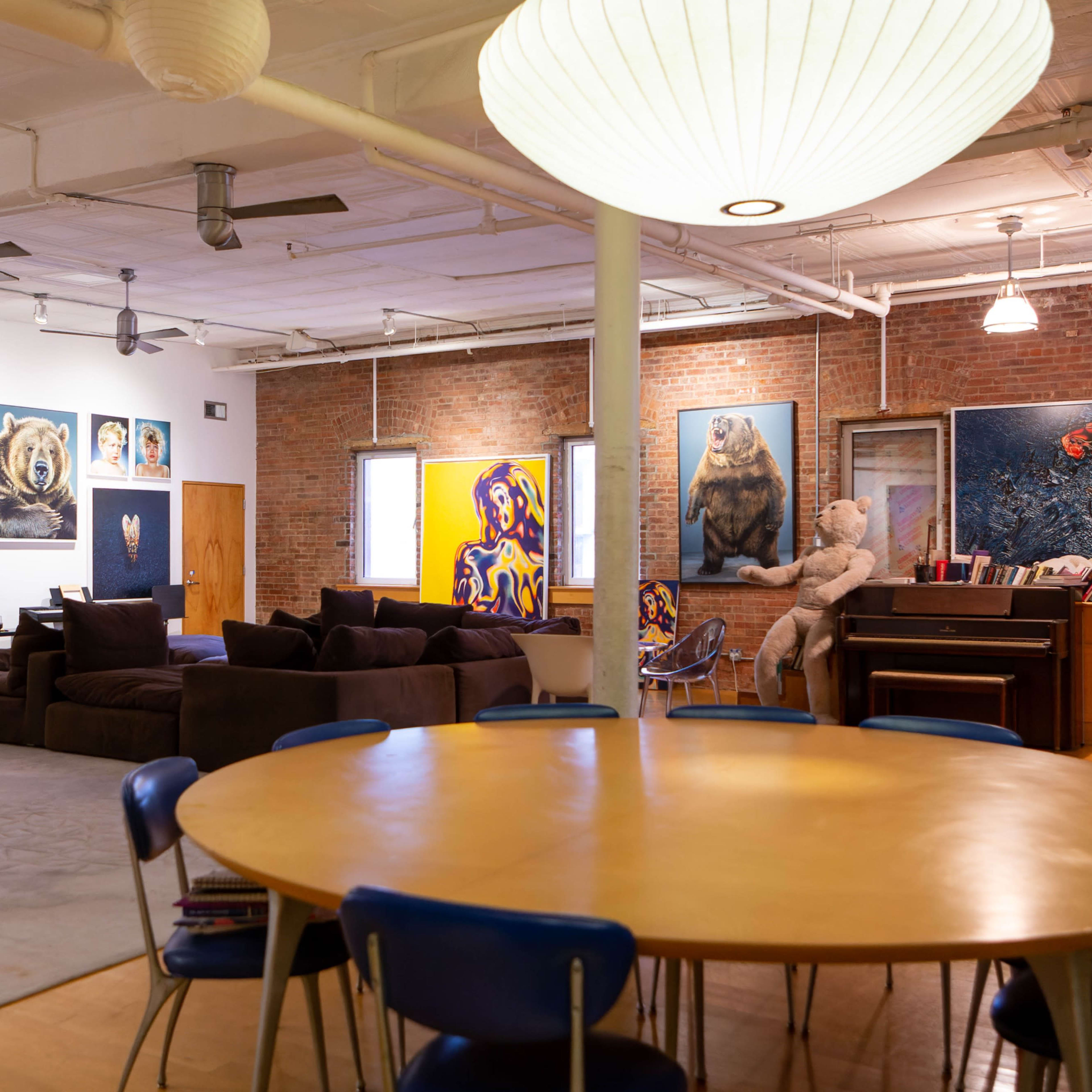 A spacious, modern living area with a large circular wooden dining table, a sectional sofa, various artworks on the walls, and a piano against a wooden panel.