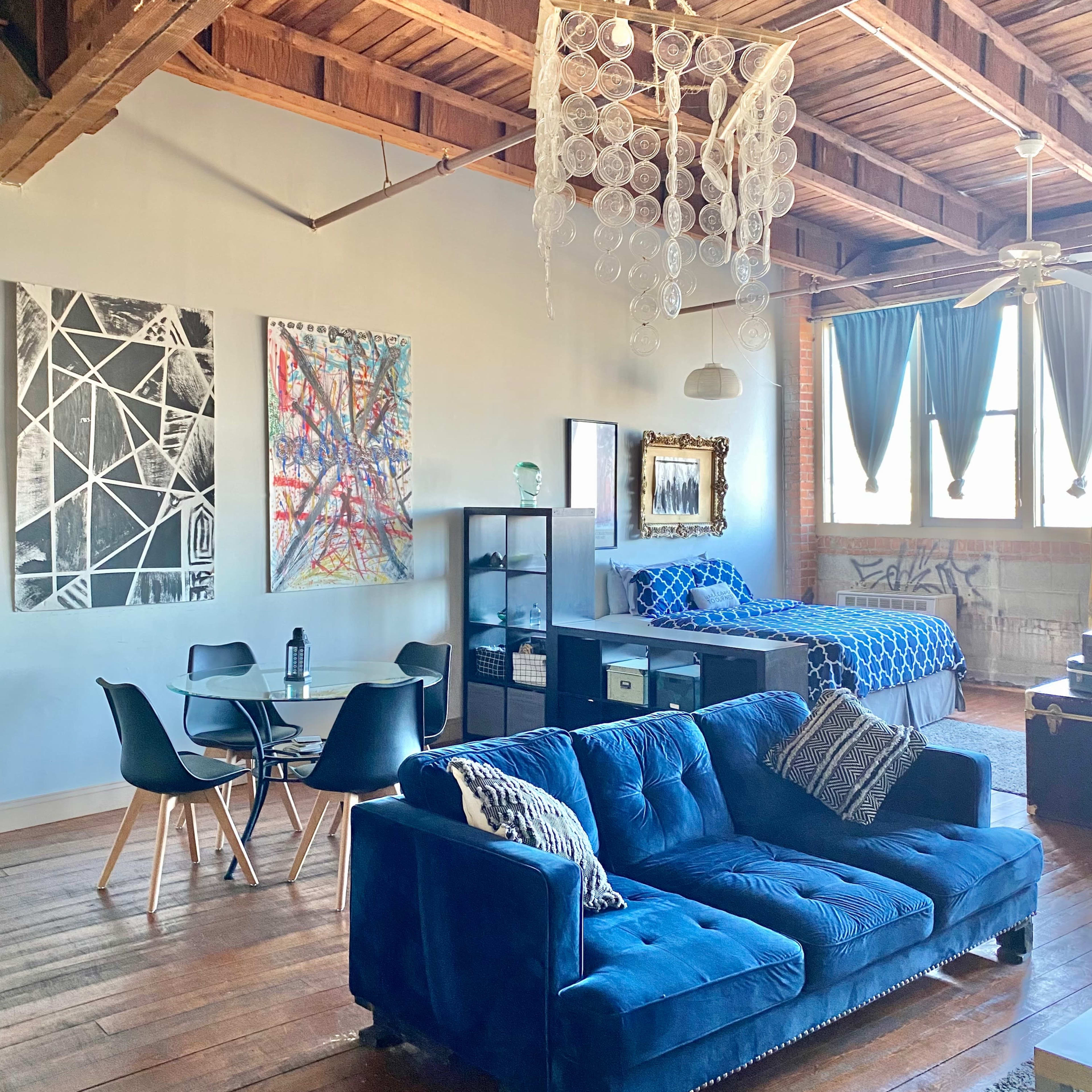 A spacious loft with wooden beams, featuring a blue couch, a circular dining table, and a bed area adorned with blue bedding.