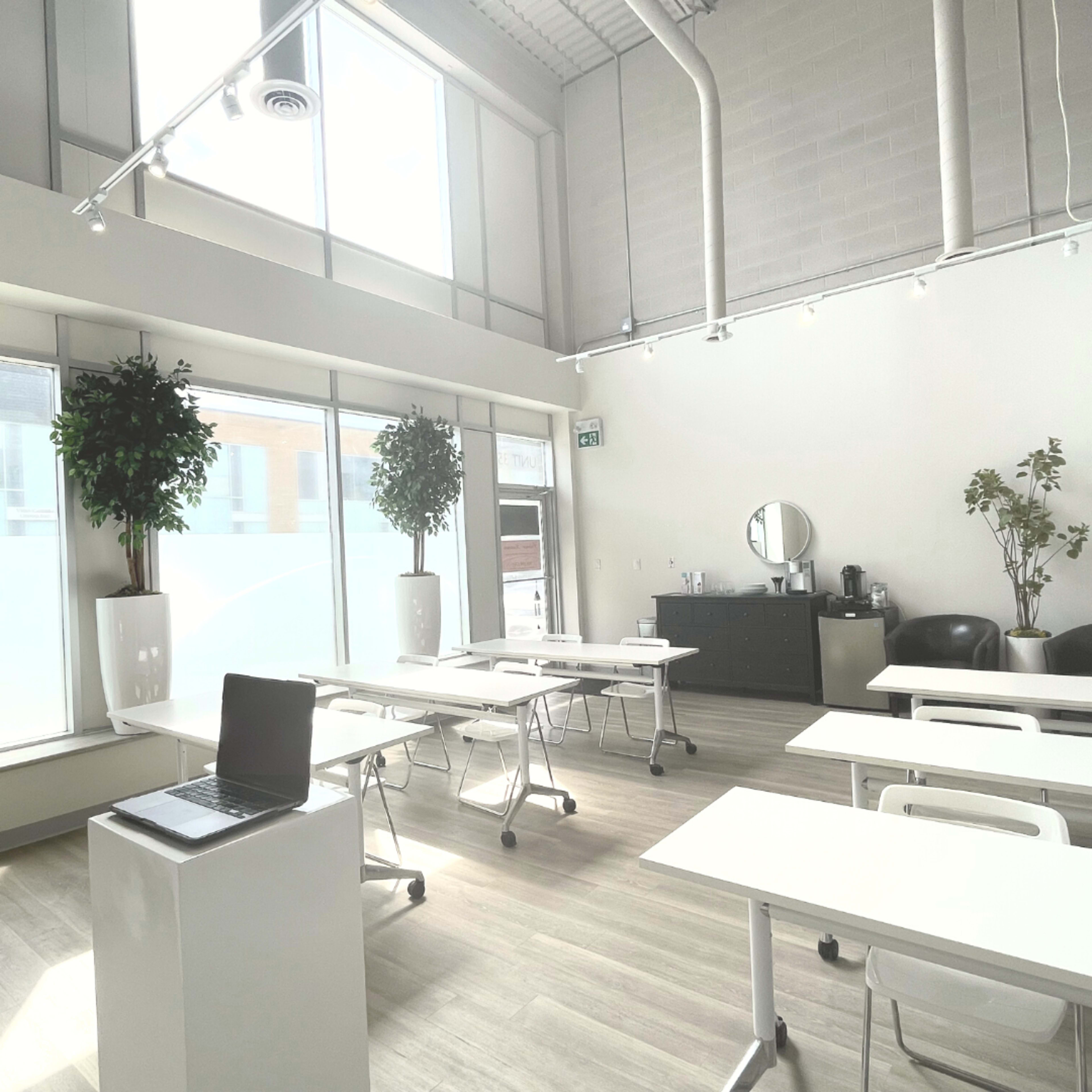 A bright, modern classroom equipped with white tables, chairs, and a large window with greenery.