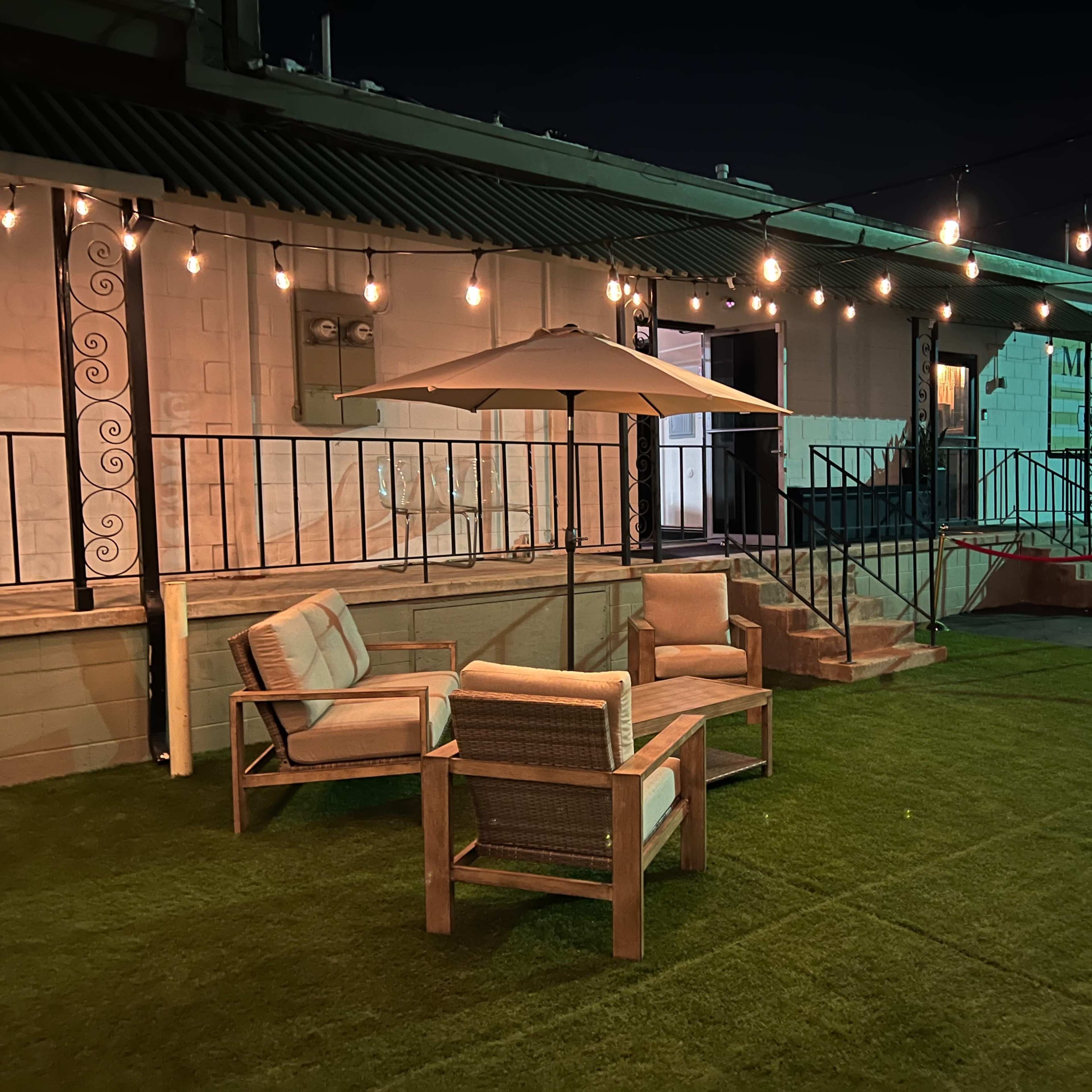 A seating area with two armchairs and an umbrella on a grassy surface, illuminated by string lights against a building at night.