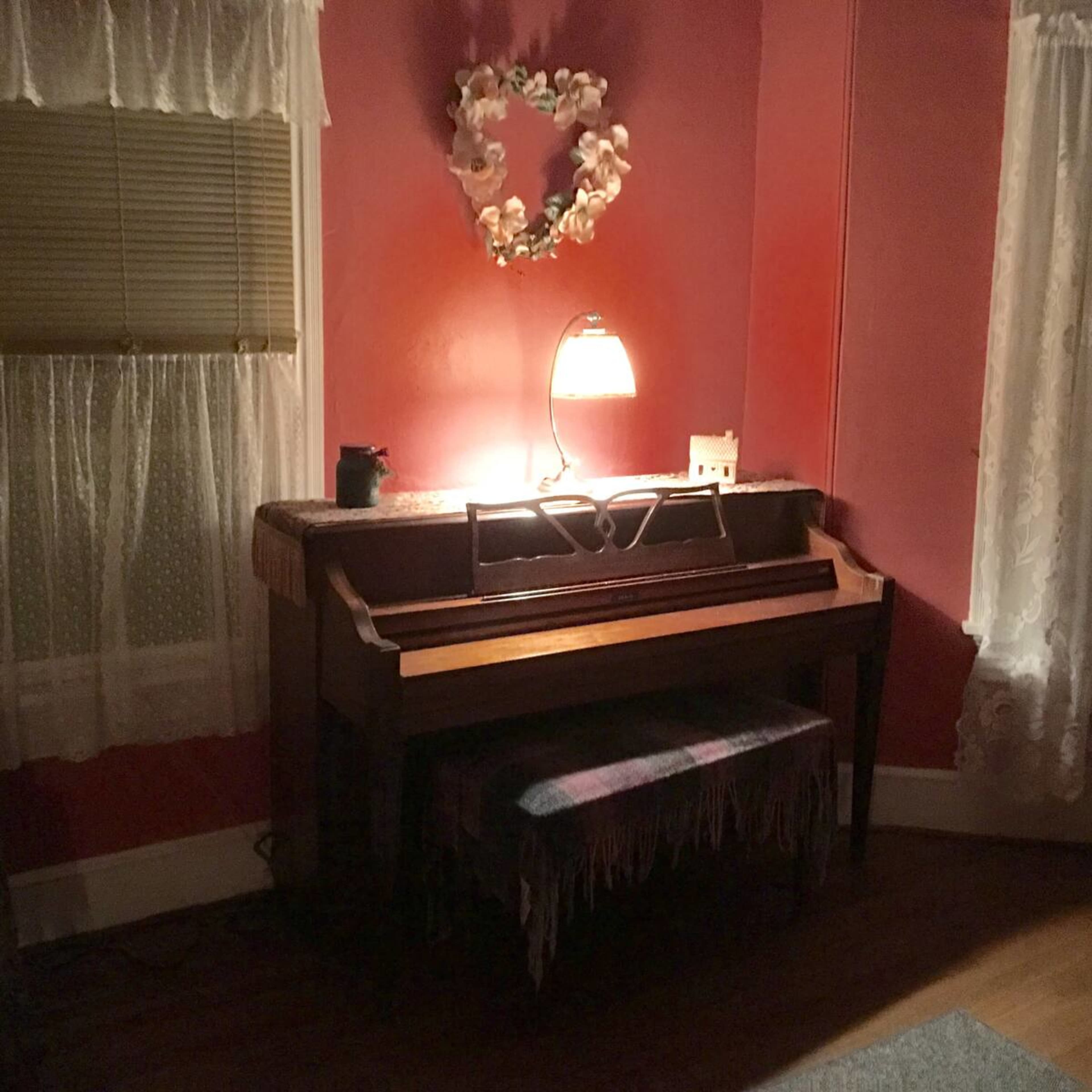 A wooden piano sits in a dimly lit corner of a room, accompanied by a small lamp and a decorative wreath on the wall.