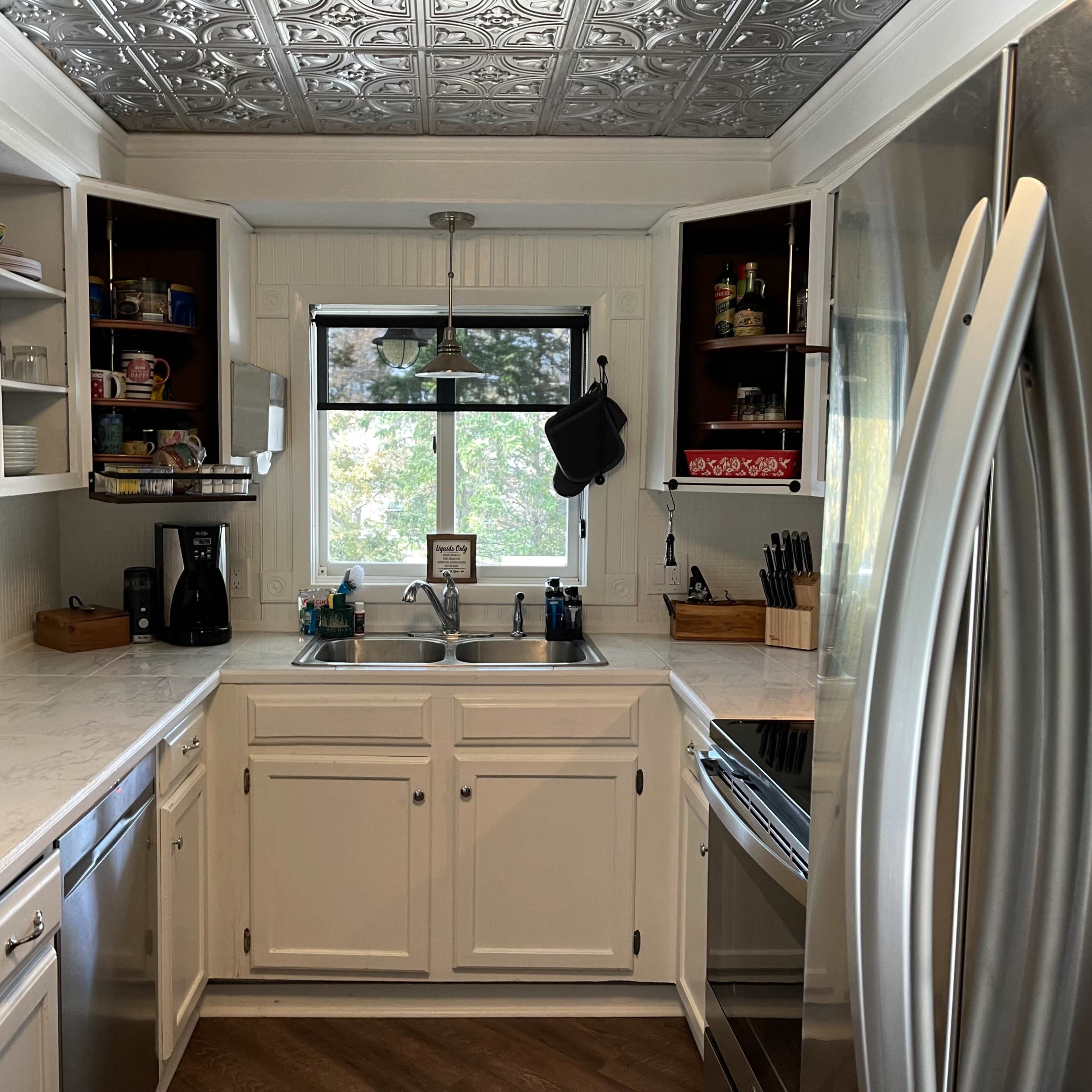 A bright kitchen with white cabinets, a marble countertop, and stainless steel appliances, featuring a window with a view of greenery outside.