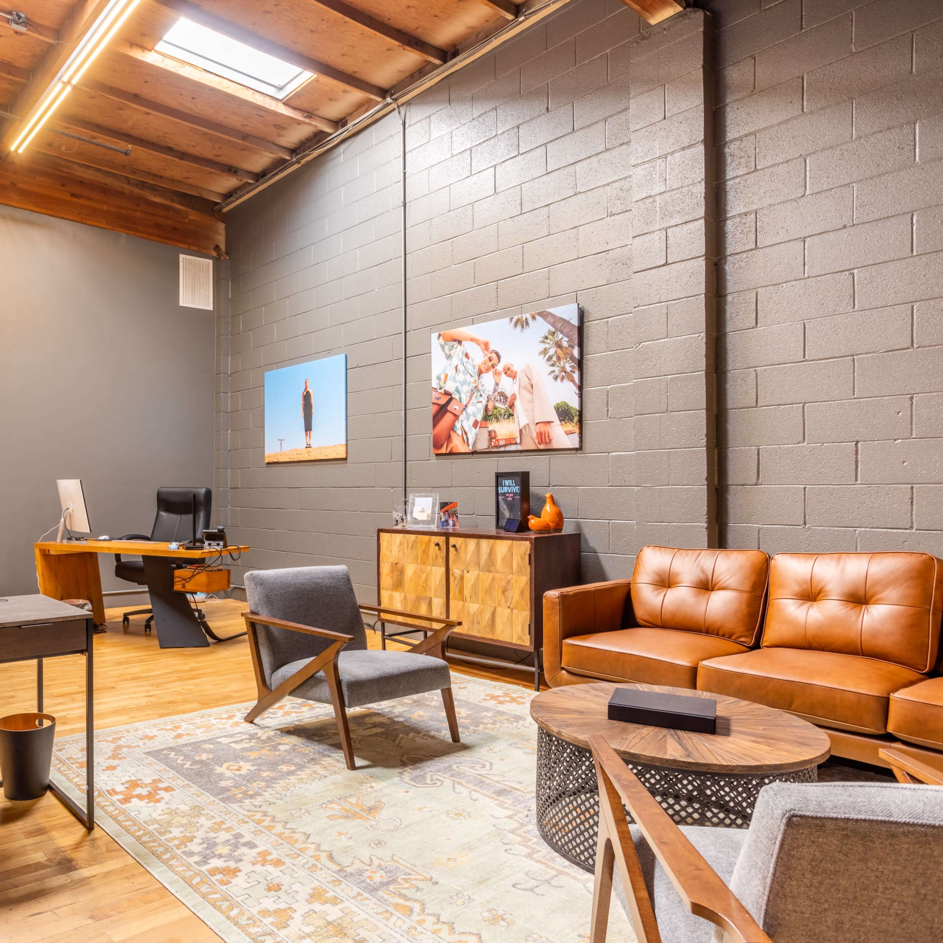 A modern office space featuring a wooden desk and chairs, a leather sofa, a large window with natural light, and decorative wall art.