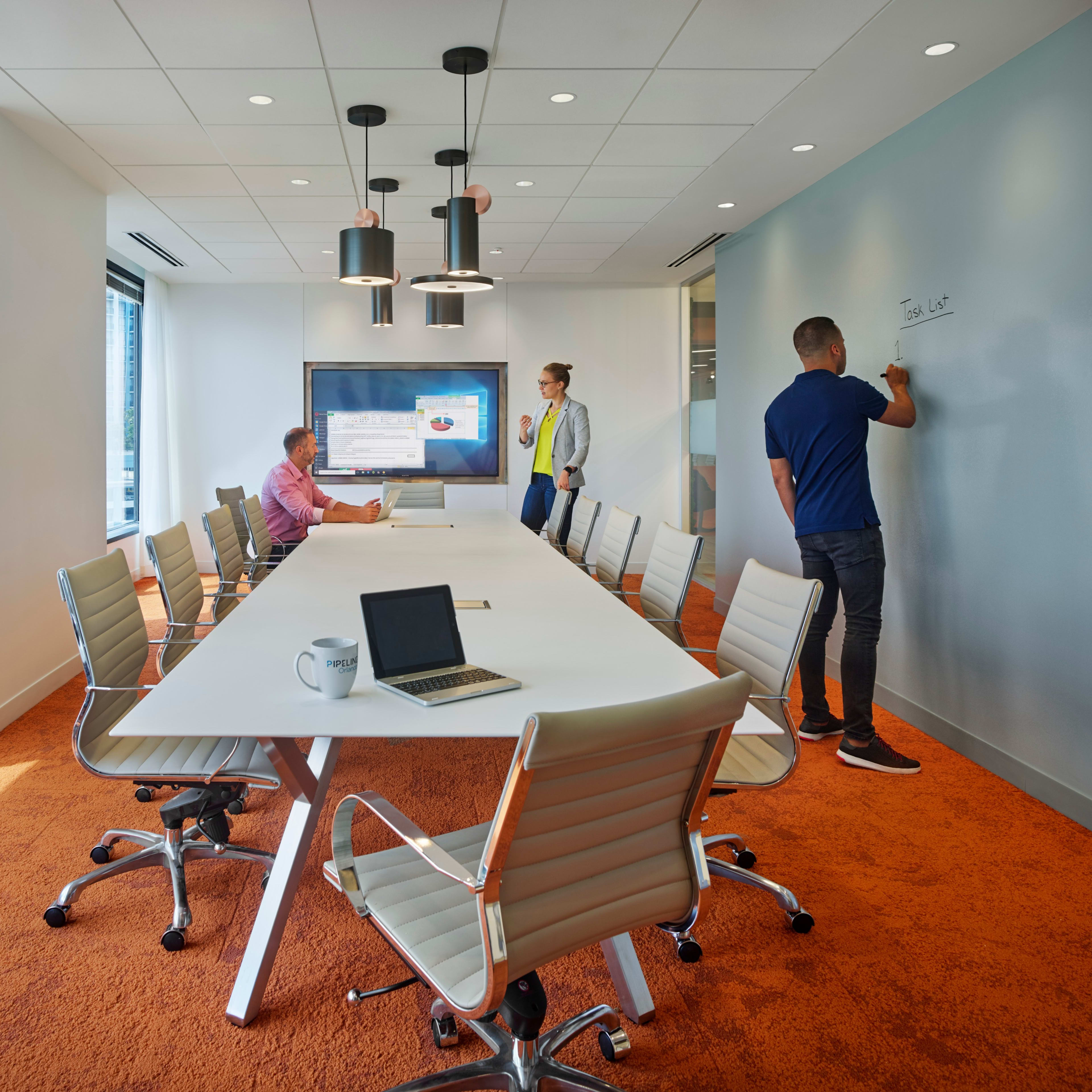A modern conference room features a long table surrounded by ergonomic chairs, with a large screen displaying a presentation and two individuals writing on a wall.