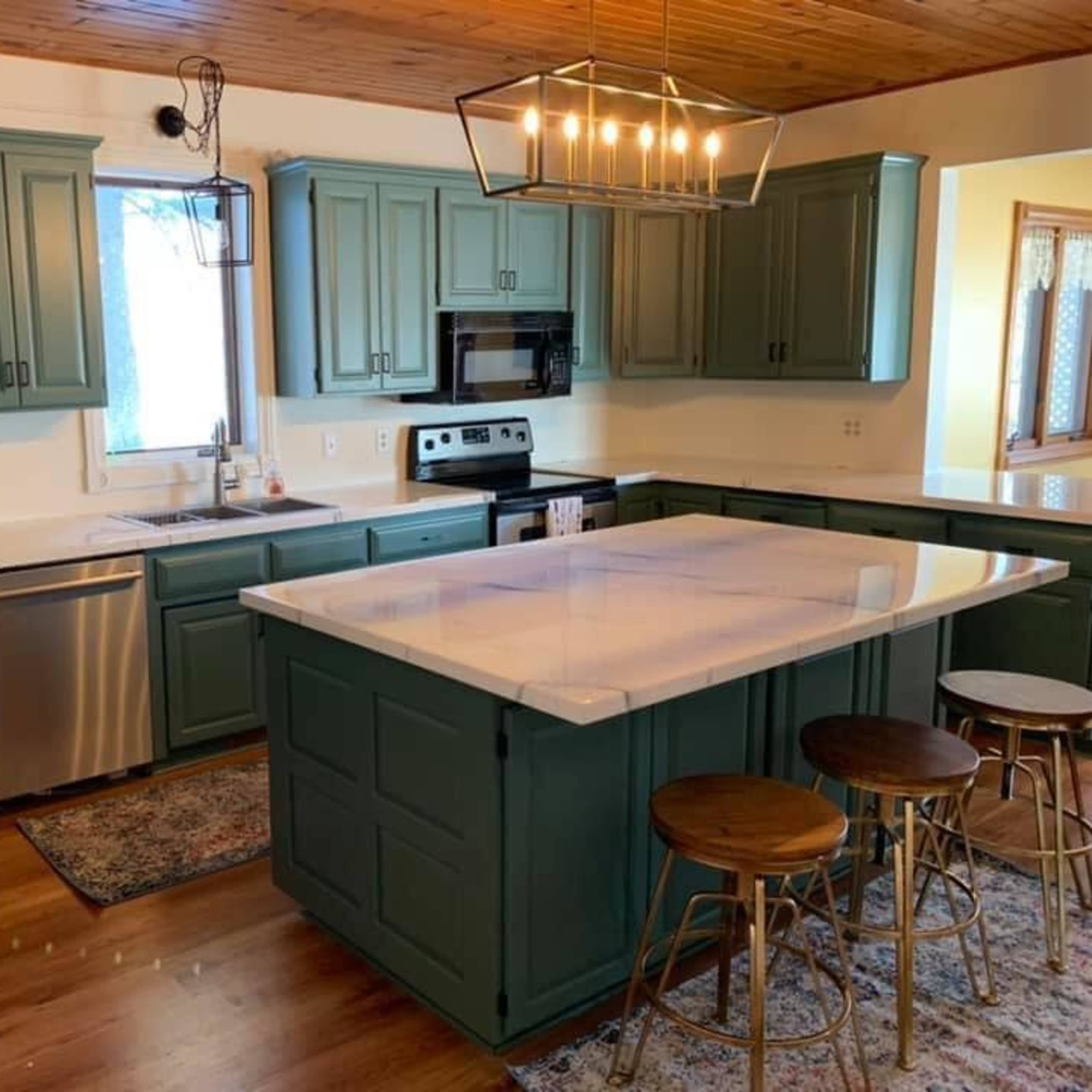 A kitchen with green cabinetry, a marble countertop island, stainless steel appliances, and wooden flooring.