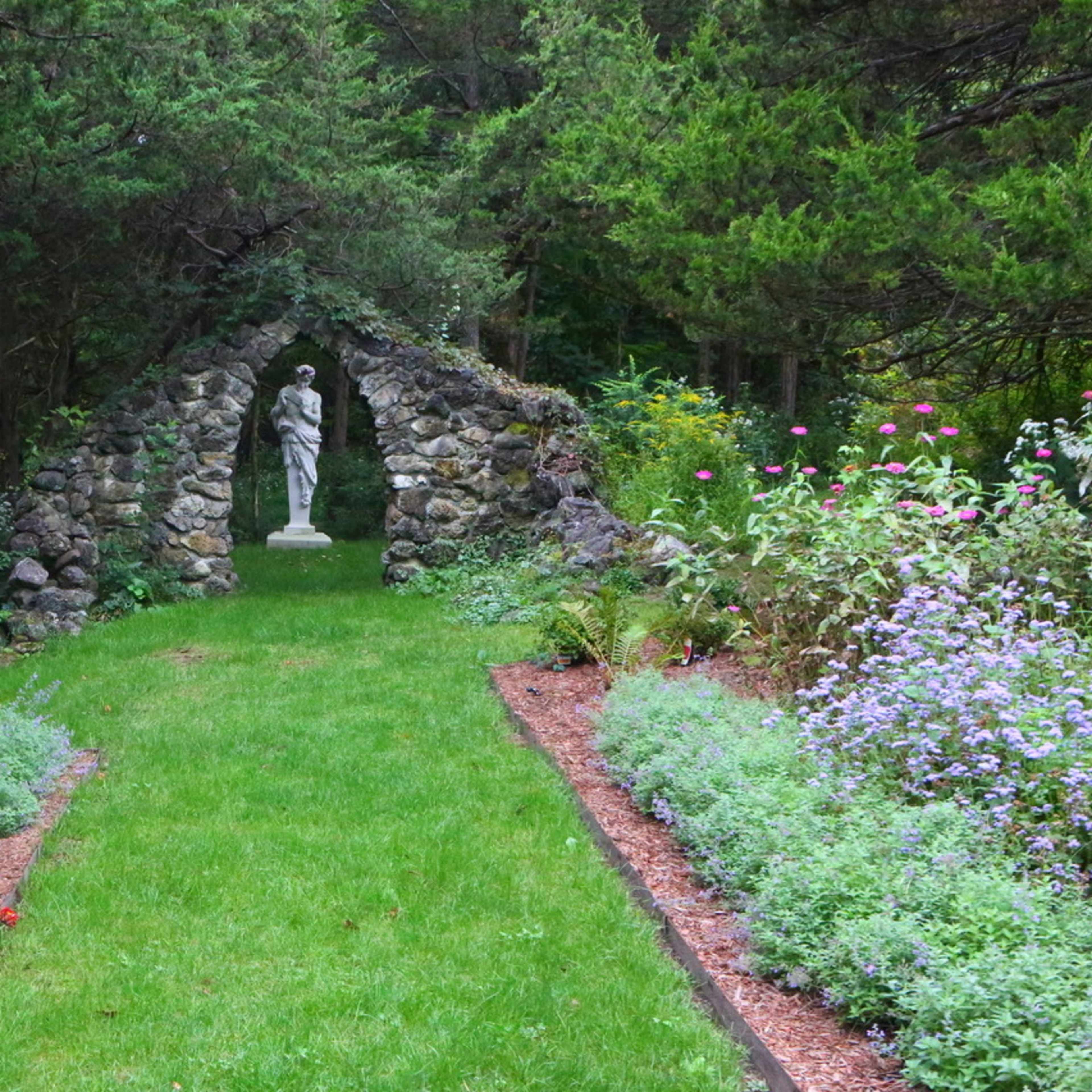 A stone archway frames a statue at the end of a grassy path lined with colorful flowers.