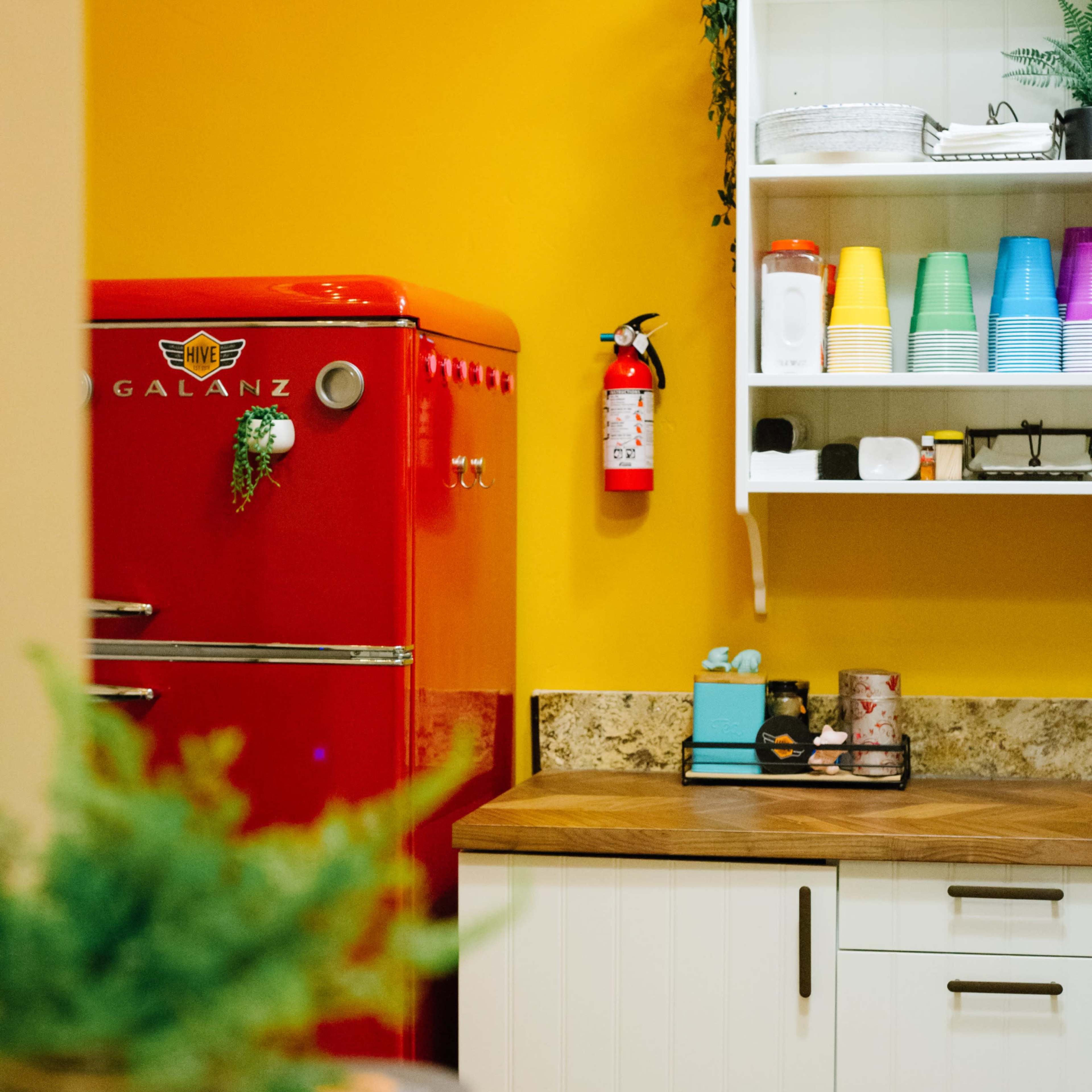 A bright yellow kitchen features a red refrigerator, white shelves with colorful cups, and a wooden countertop with various kitchen items.