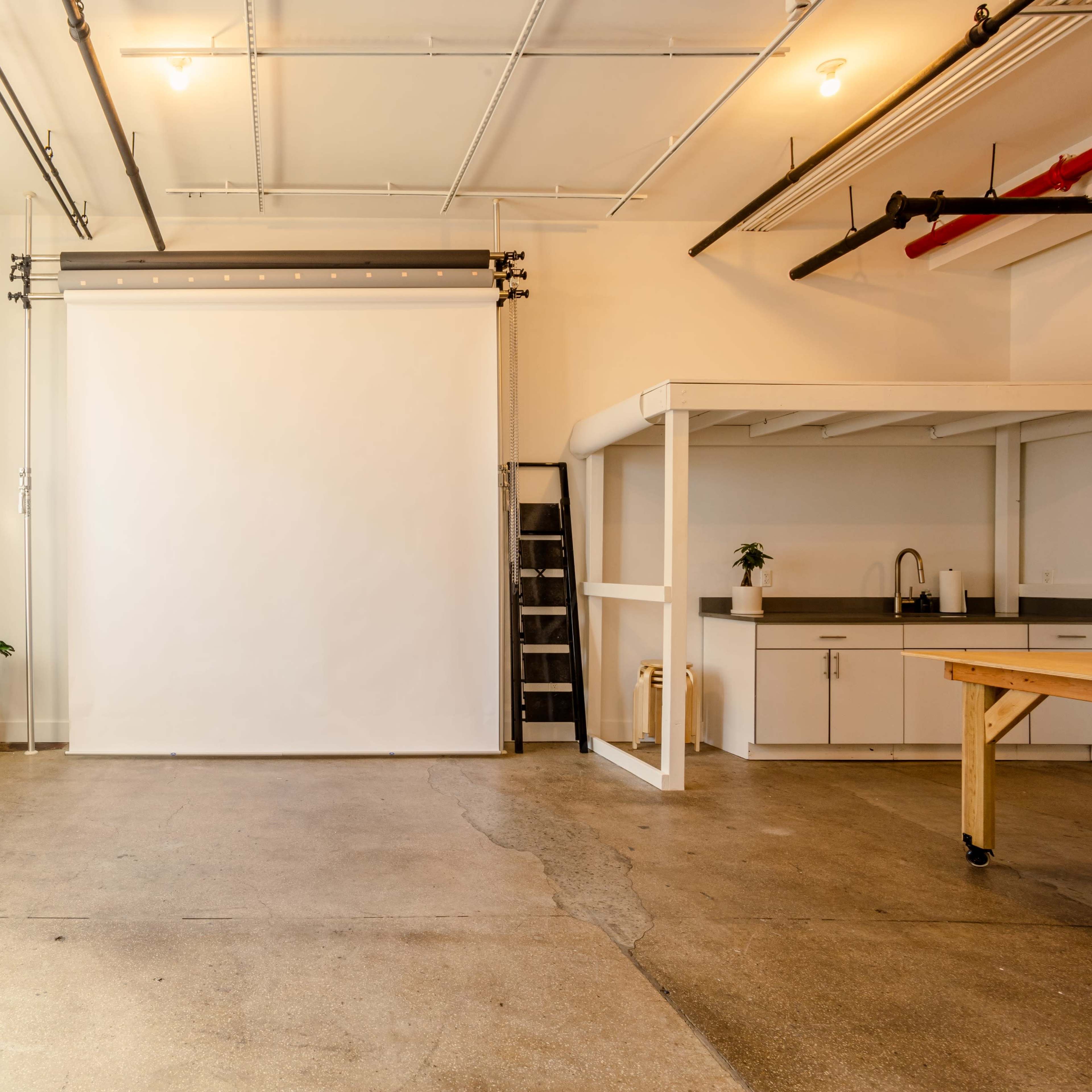 A bright, open studio space featuring a blank white backdrop, a kitchen area, and a ladder against a wall.
