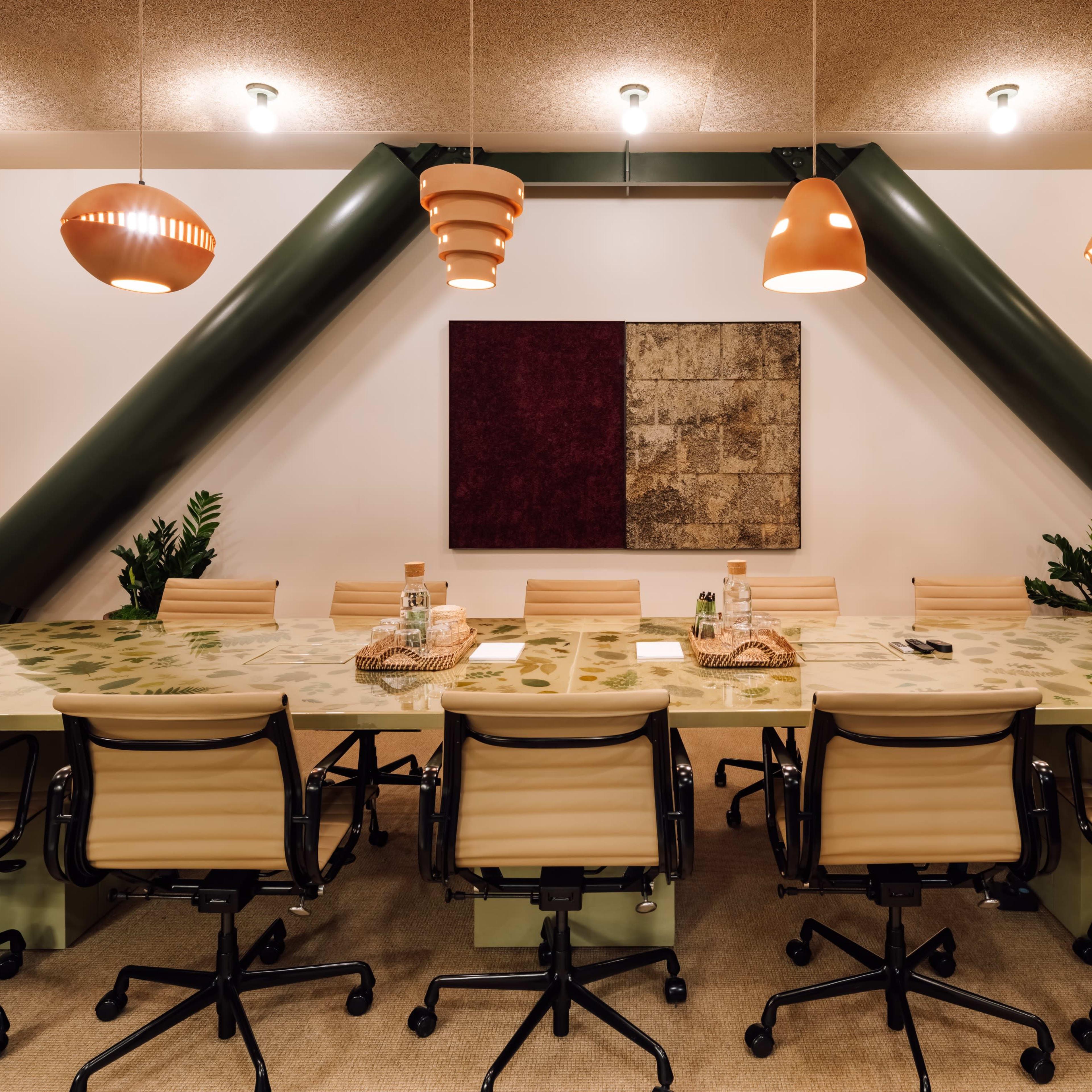A large conference room features a long table surrounded by beige chairs, with green support beams and pendant lights overhead.
