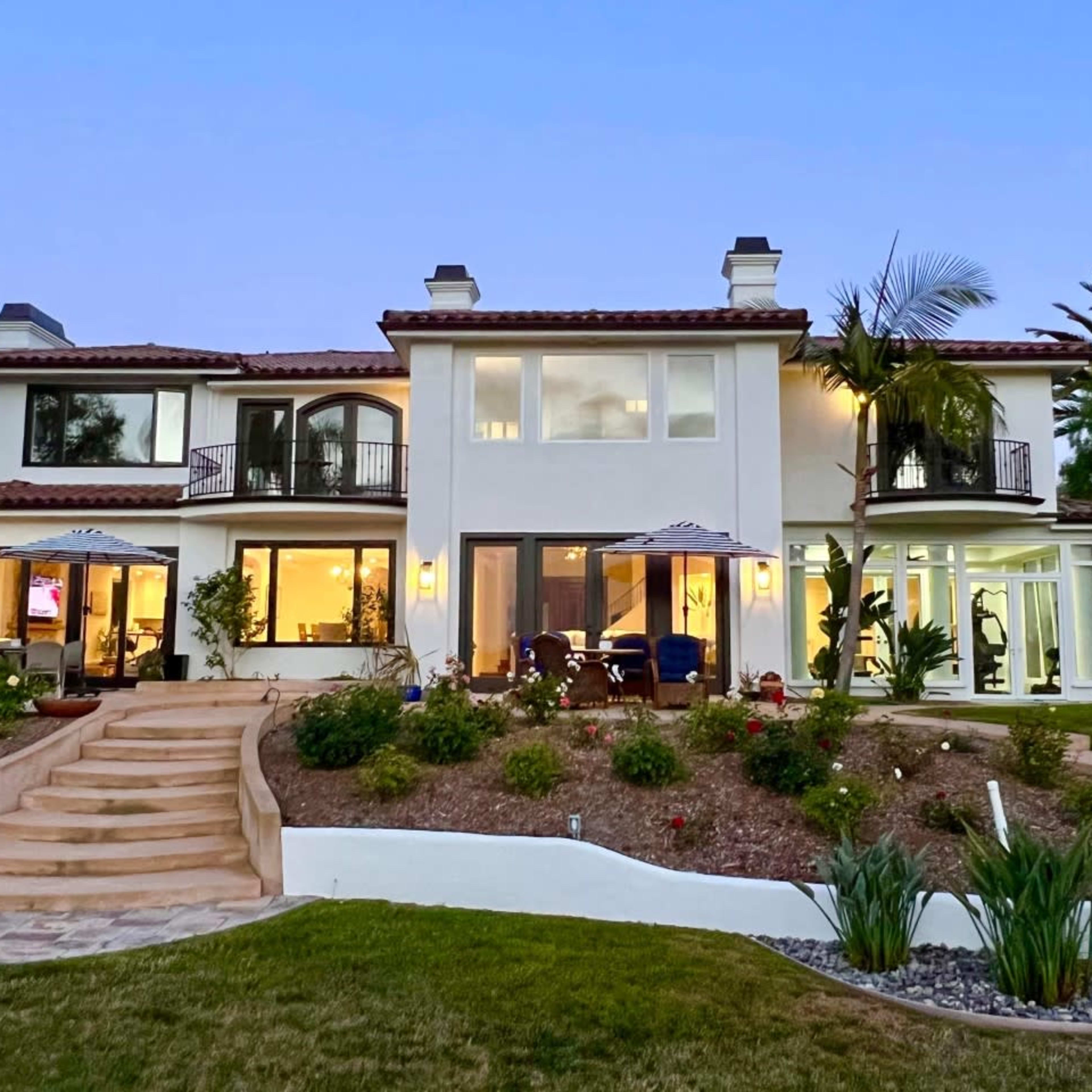 A large, two-story house with a landscaped yard and multiple balconies, illuminated by evening lights.