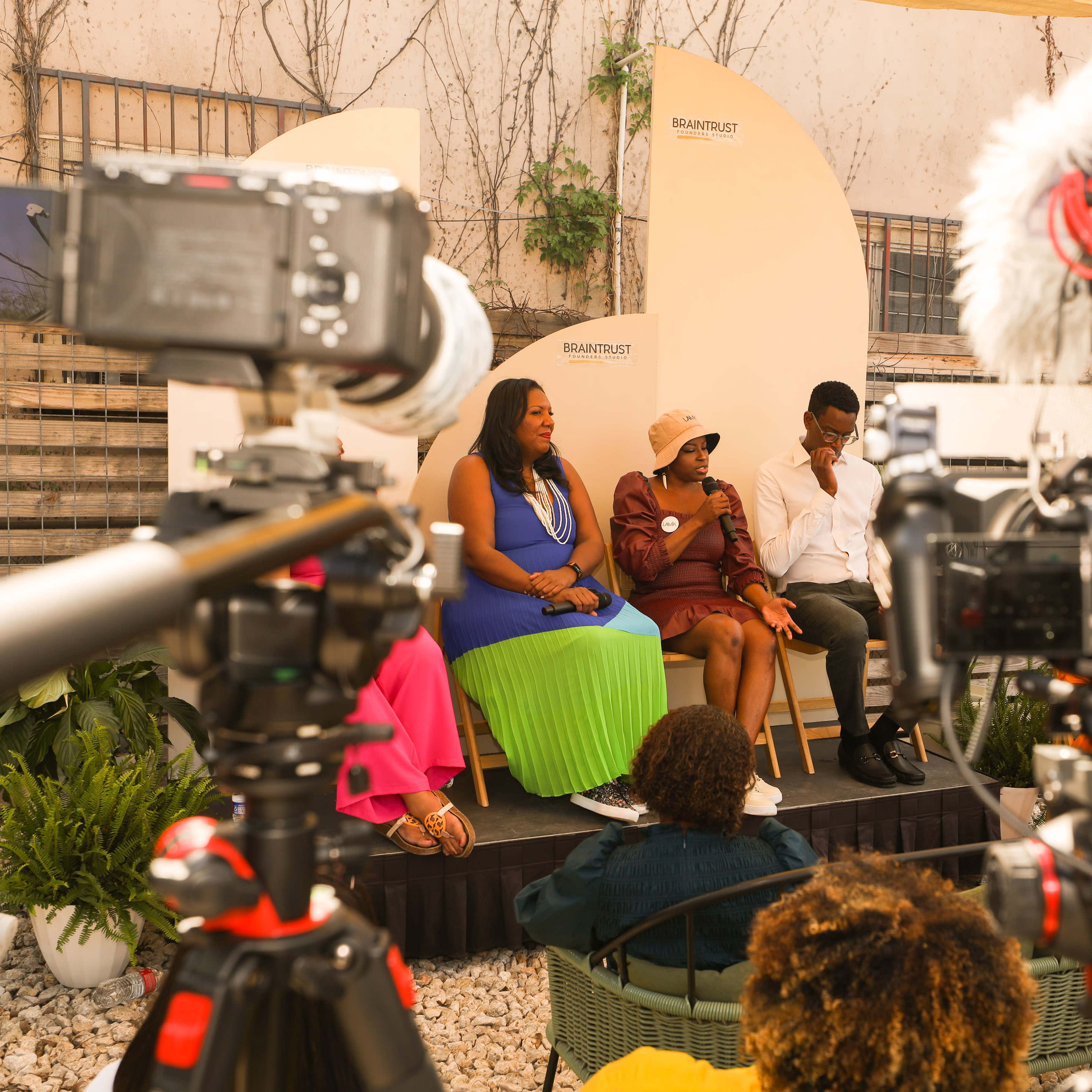 Three speakers sit on a stage during a panel discussion, with cameras focused on them in a garden setting.