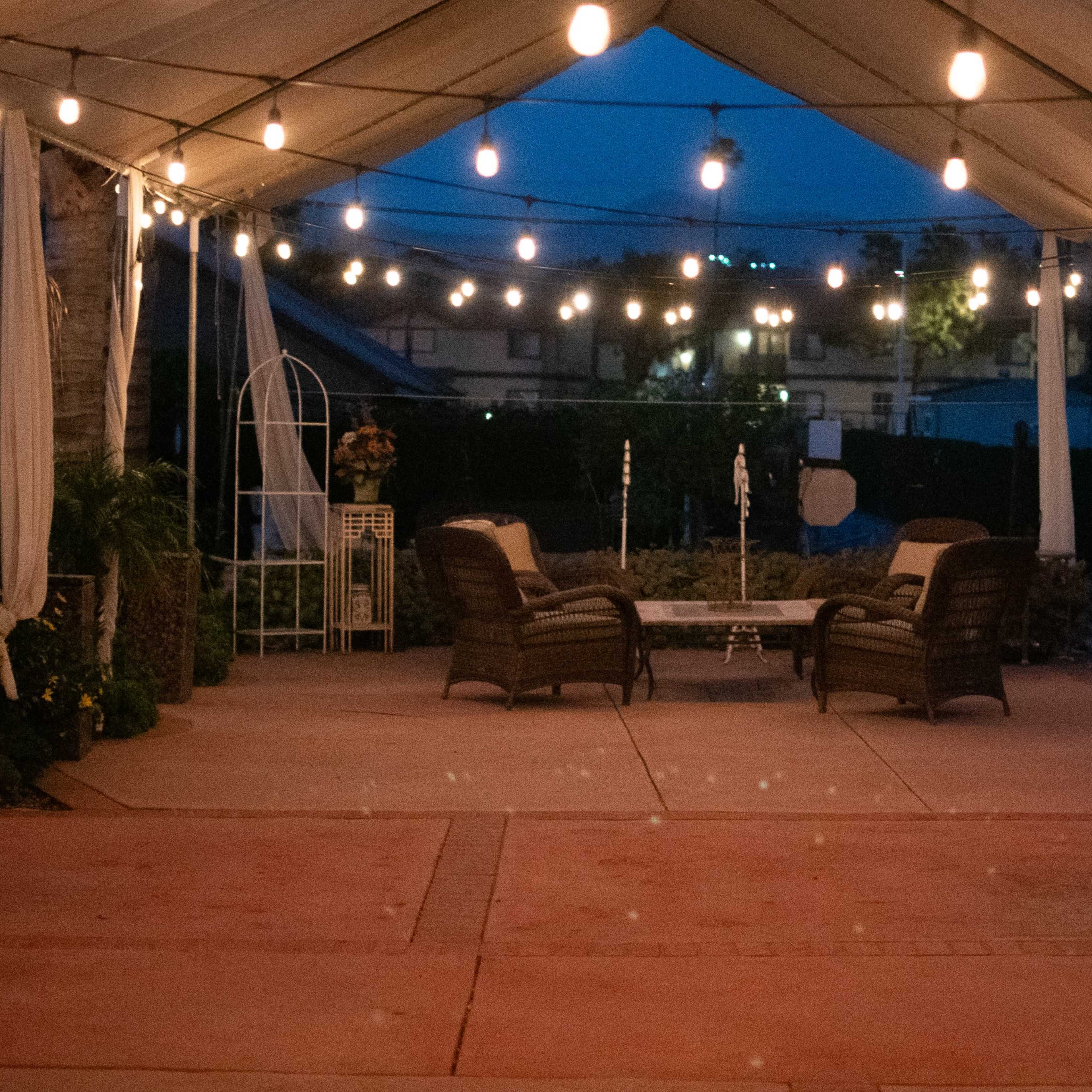 A tented outdoor area is illuminated by string lights and features two seated chairs arranged around a central table.
