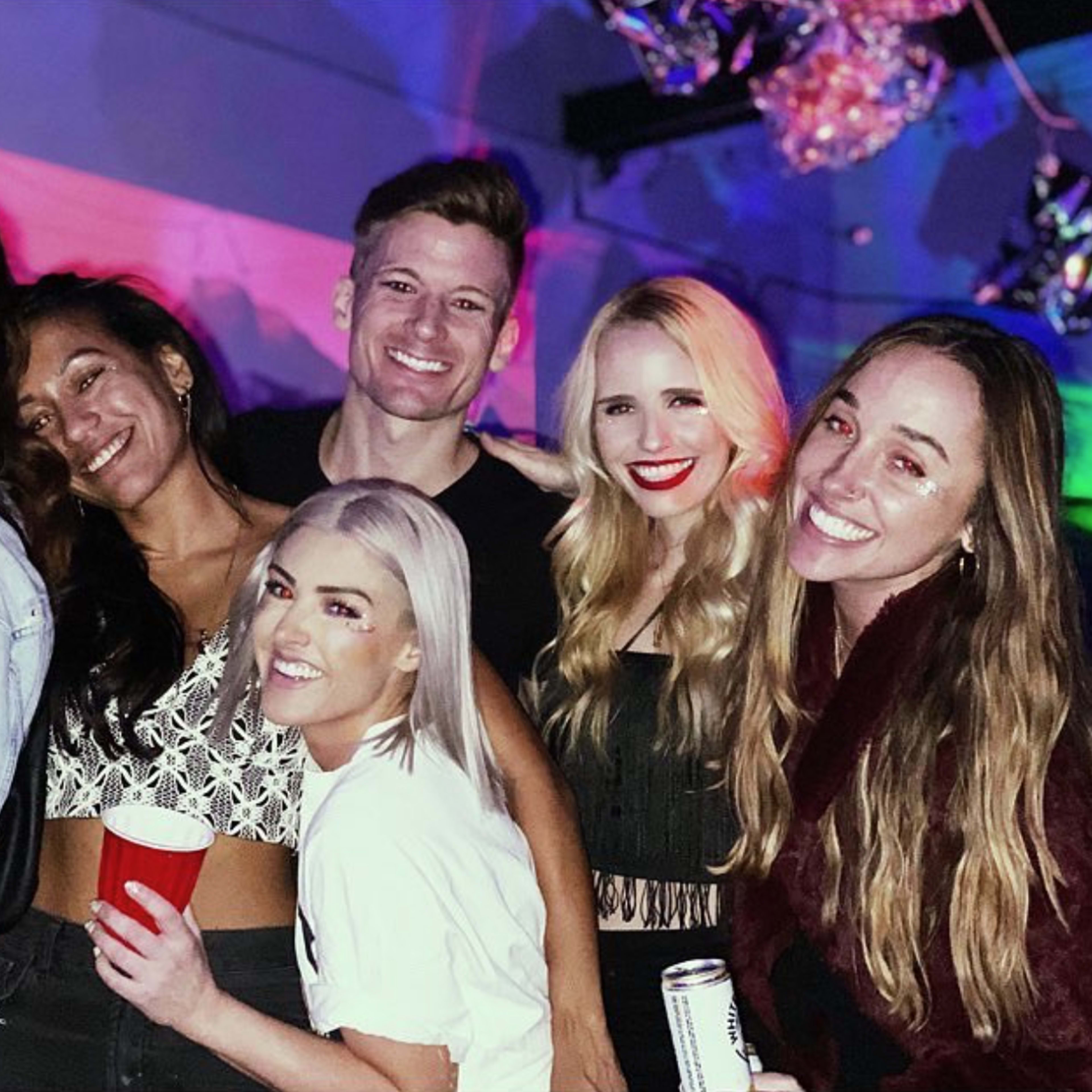 A group of six people poses closely together in a dimly lit venue decorated with colorful lights.