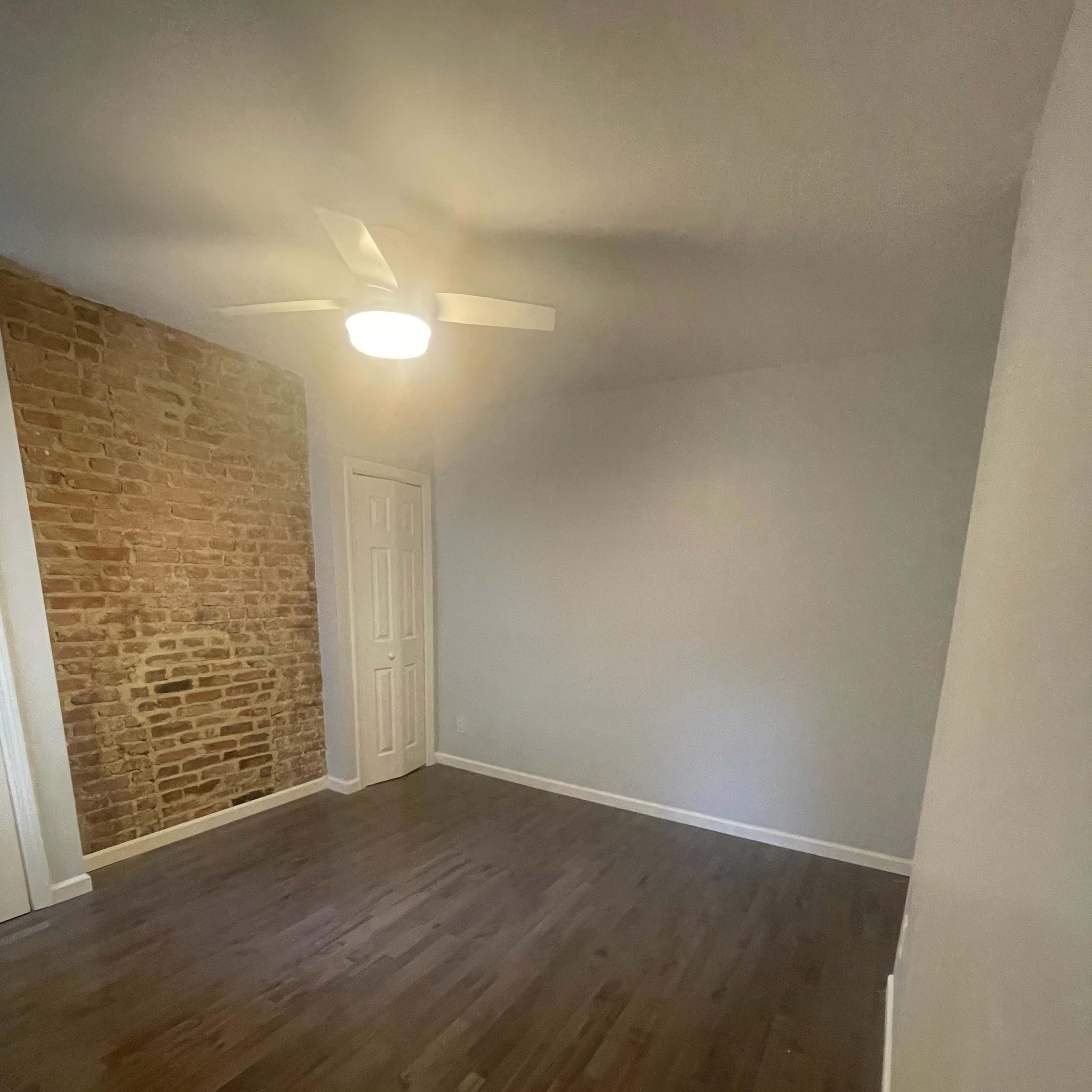 A spacious room with a wooden floor, a ceiling fan, and a brick accent wall.