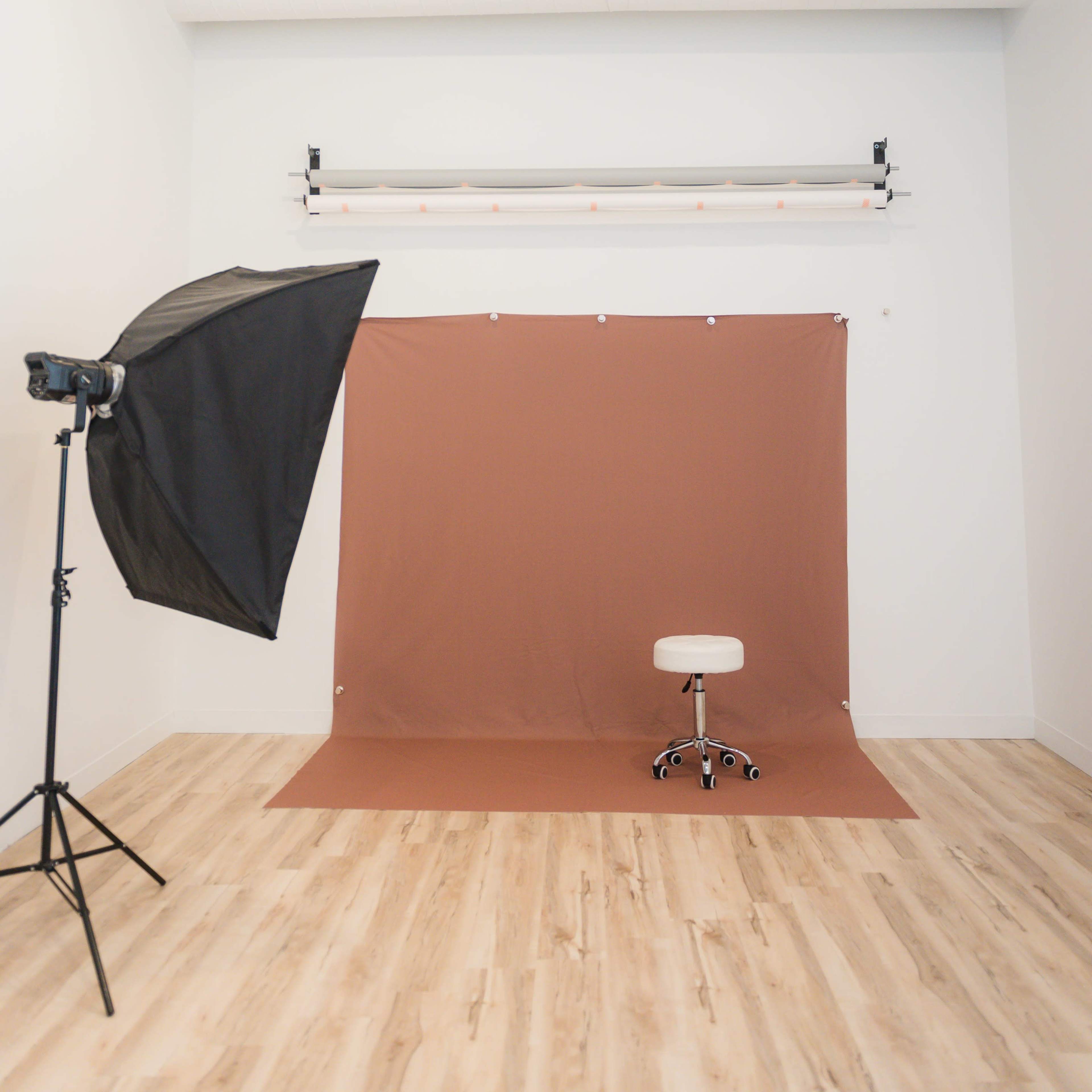 A photography studio features a brown backdrop and a white adjustable chair, illuminated by a softbox light.