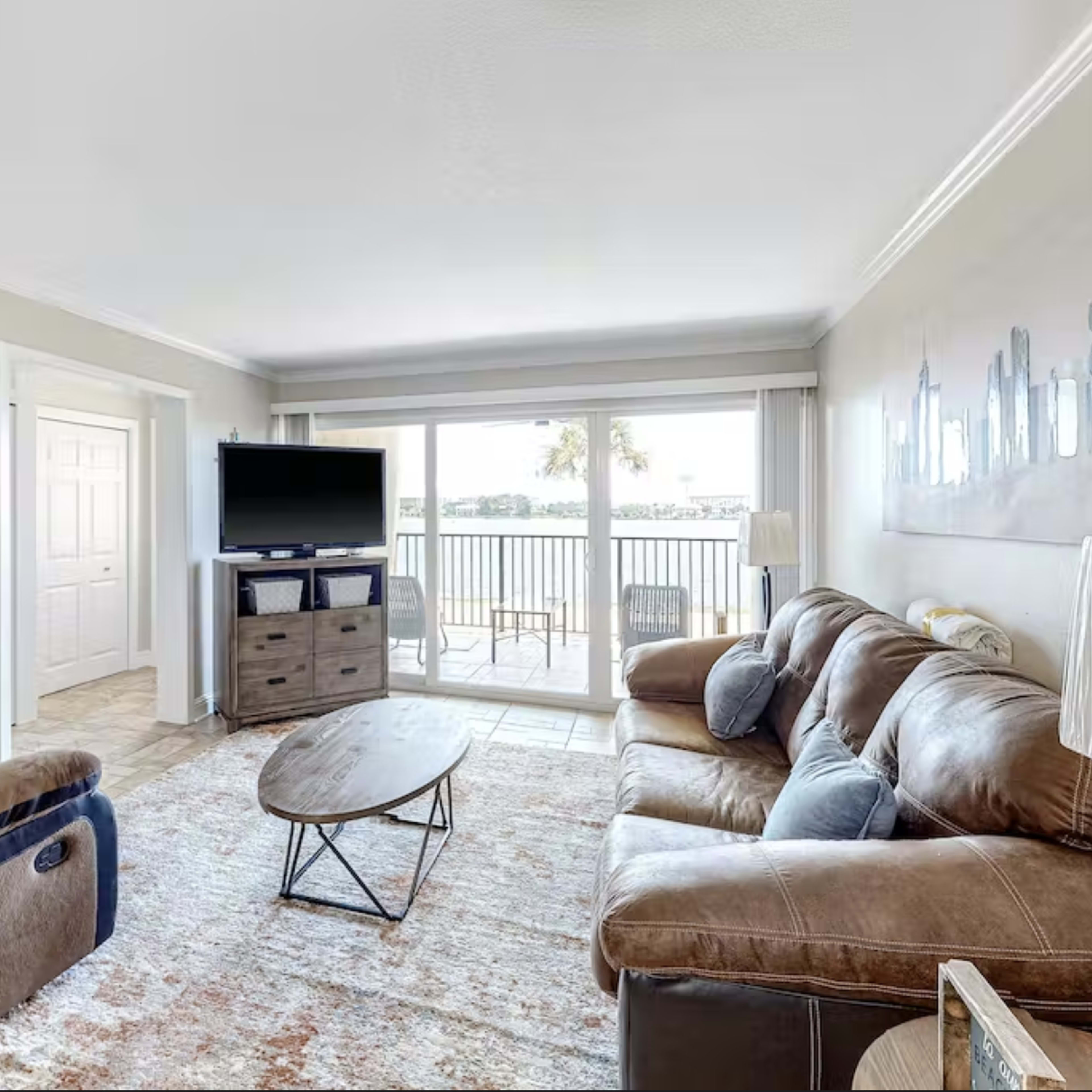 The living room features a brown leather sofa and recliner, a coffee table, a television, and large windows with a view of the outdoors.
