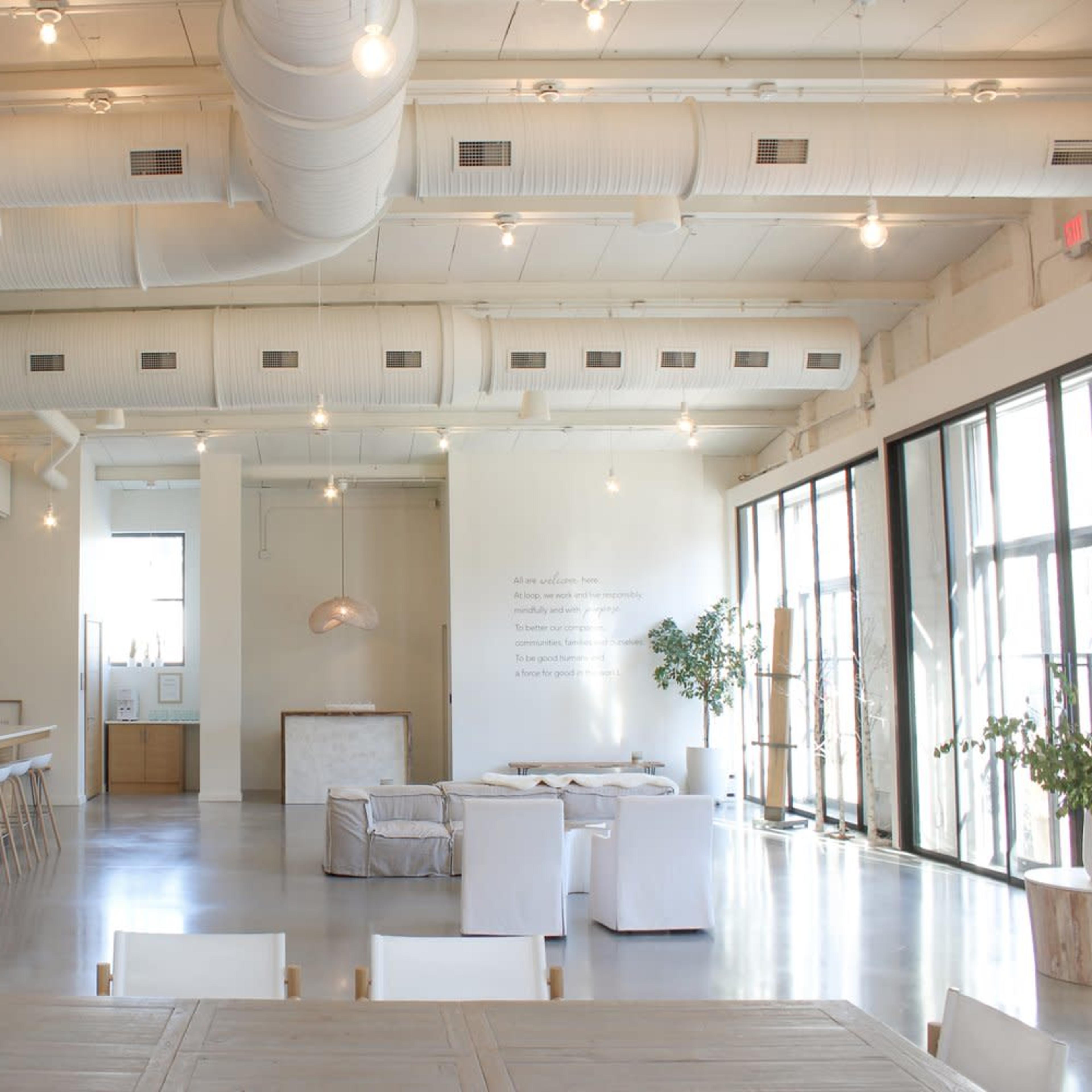 A spacious, modern interior with high ceilings, large windows, and minimalist furniture arranged in a dining and lounging area.