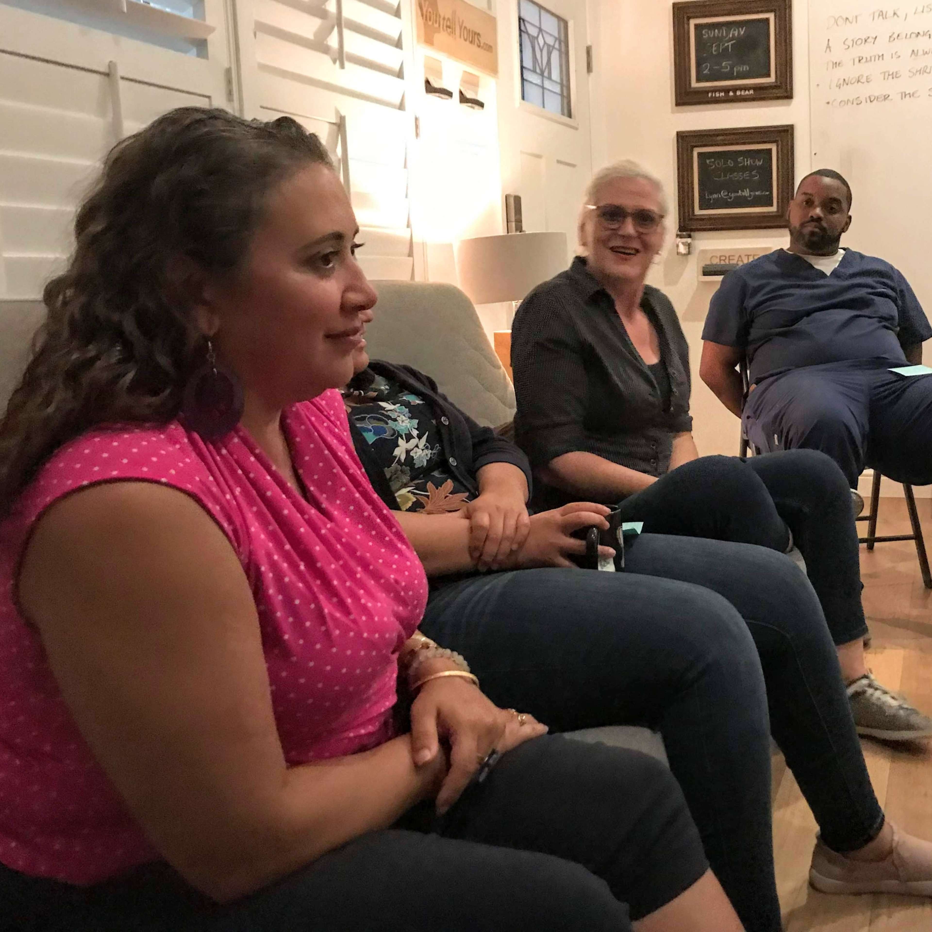 A group of people is seated in a circle in a room with white walls and various notes displayed.