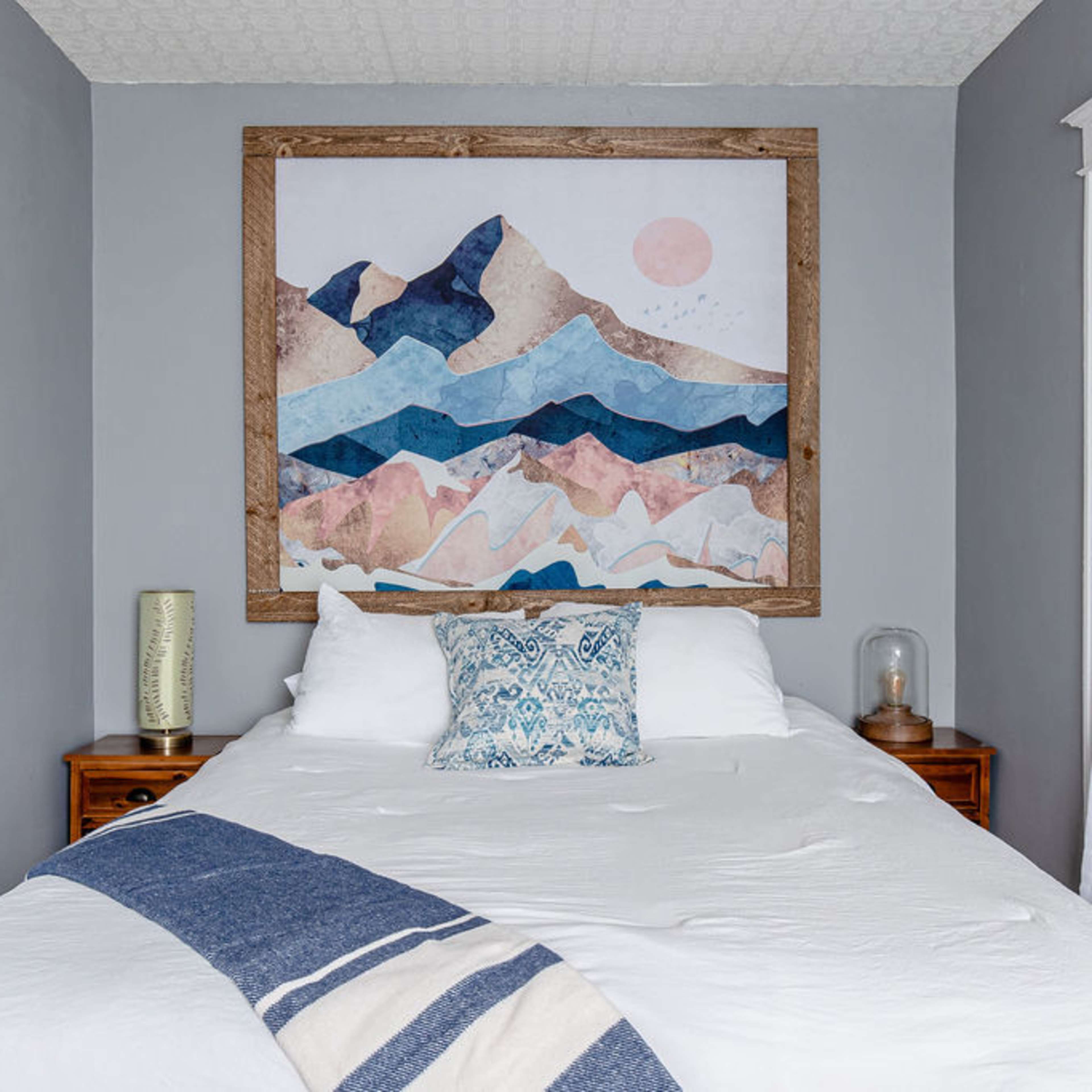 A bedroom features a bed with a white comforter, a decorative pillow, and a large mountain-themed artwork above the headboard.