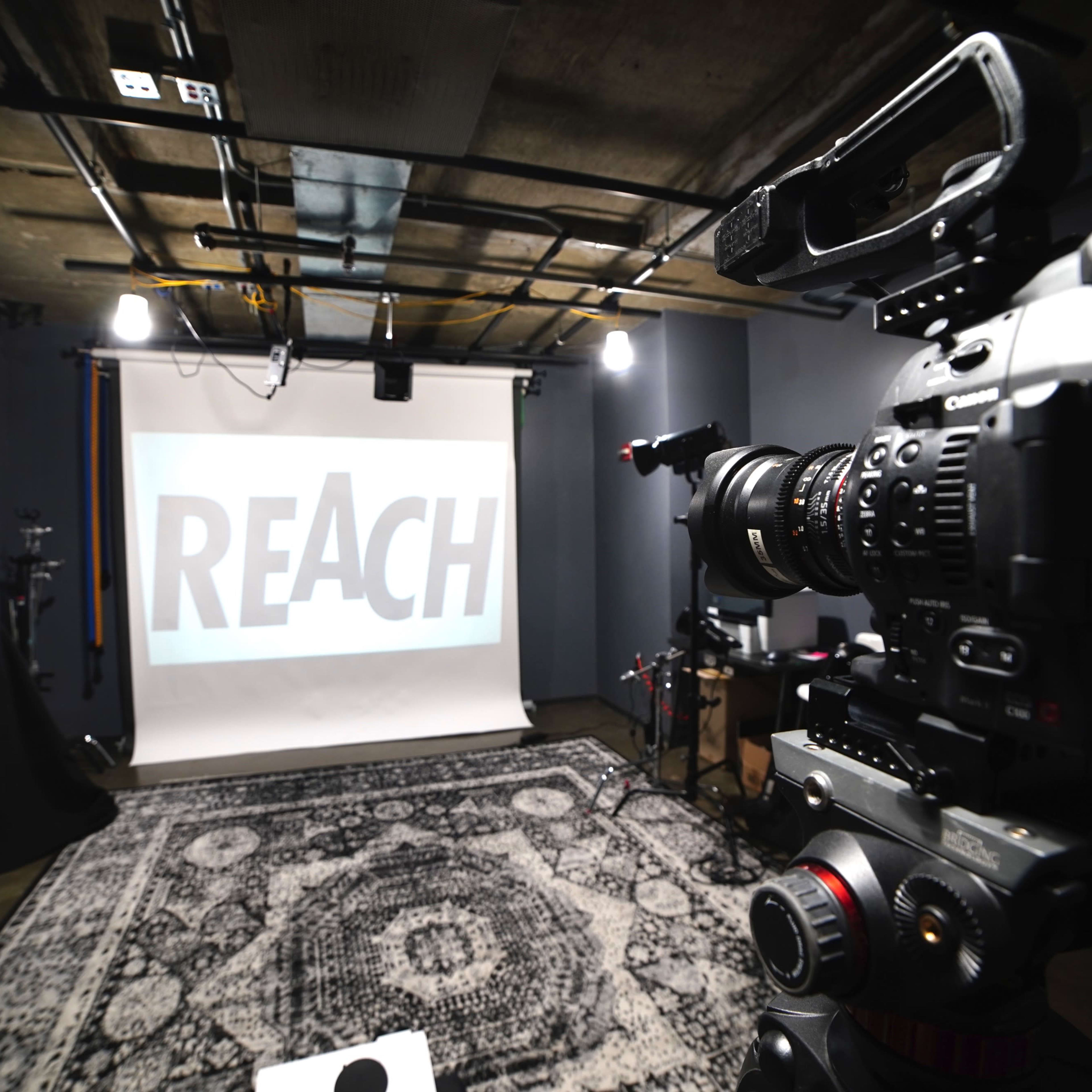 A camera is positioned in front of a projection screen displaying the word "REACH" in a studio setting with dim lighting and a patterned rug.