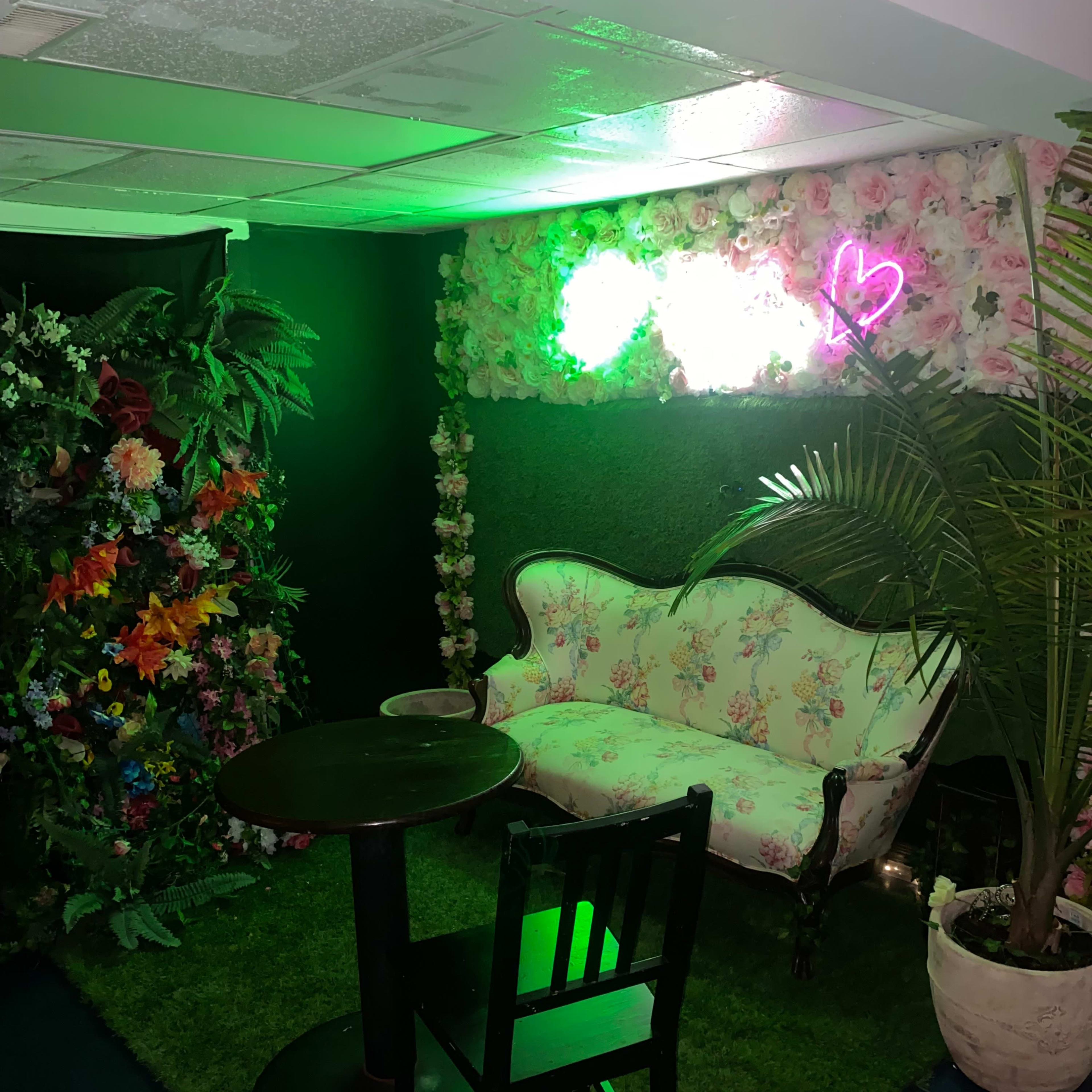 The scene features a floral-themed seating area with a vintage-style sofa, table, and greenery, set against a backdrop of artificial plants and neon lights.