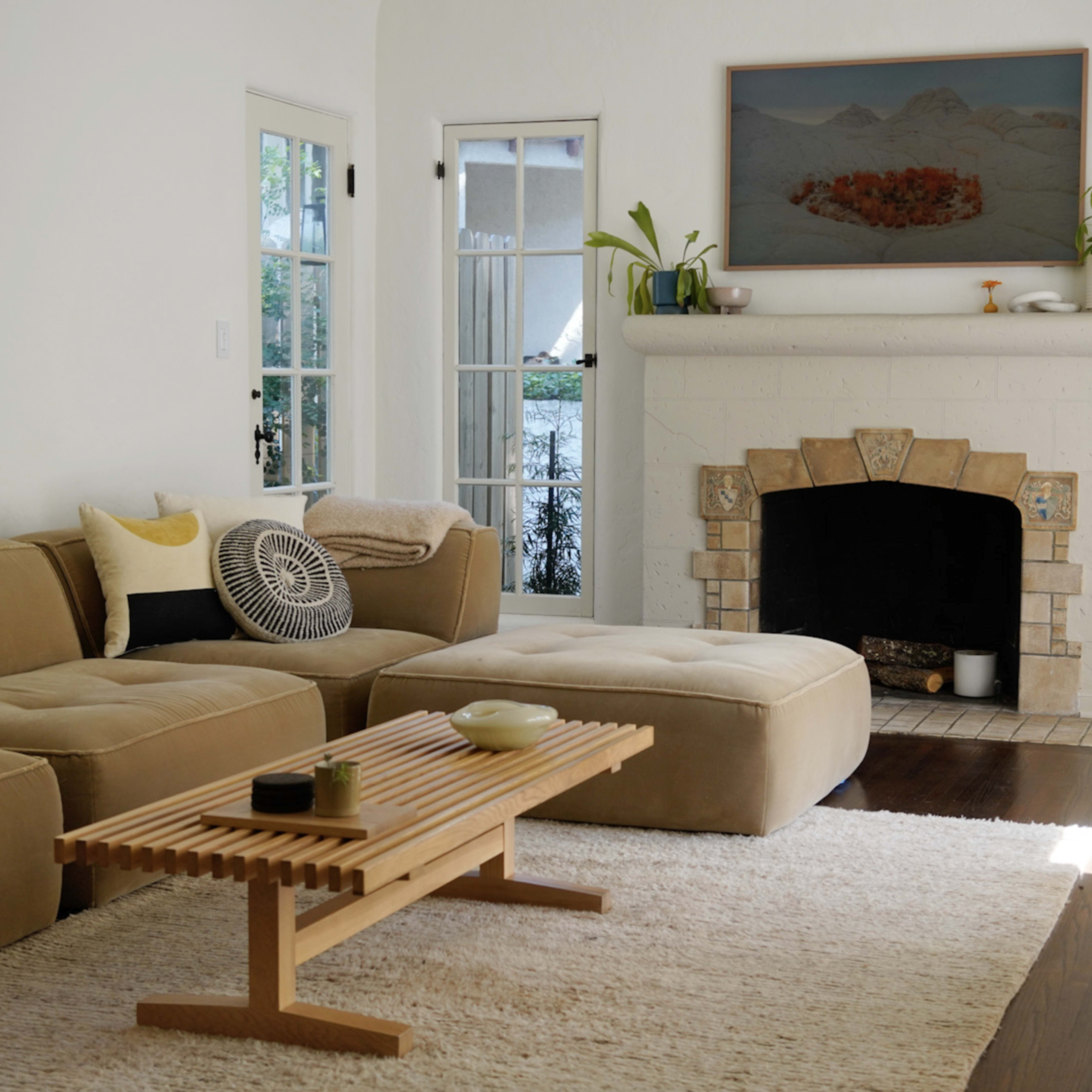 A living room with a beige sectional sofa, a wooden coffee table, a rug, and a fireplace surrounded by light-colored walls and large windows.