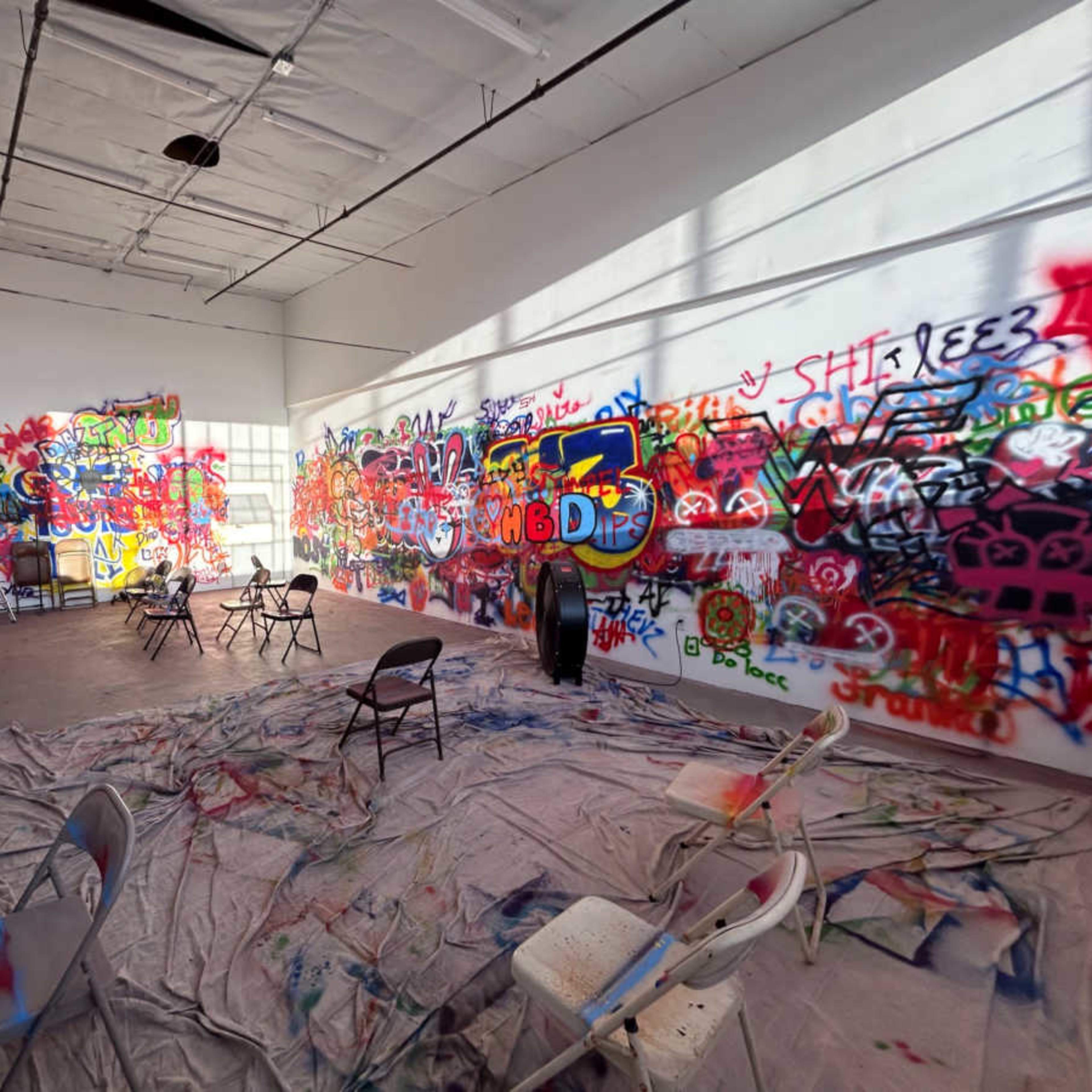 An art studio filled with colorful graffiti on white walls, chairs scattered around, and a tarp covering the floor.