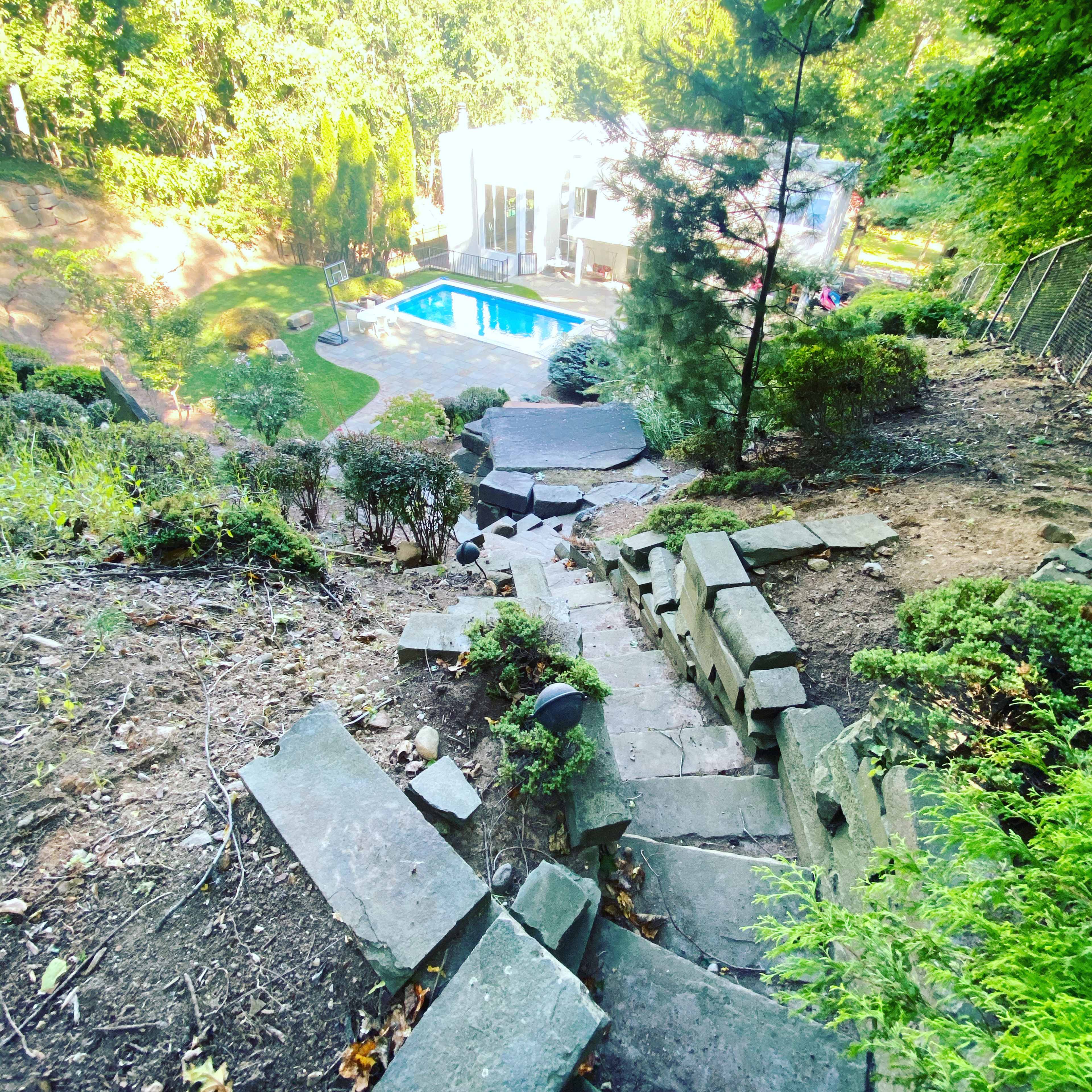 A stone staircase leading down a hillside towards a swimming pool surrounded by greenery and a house in the background.