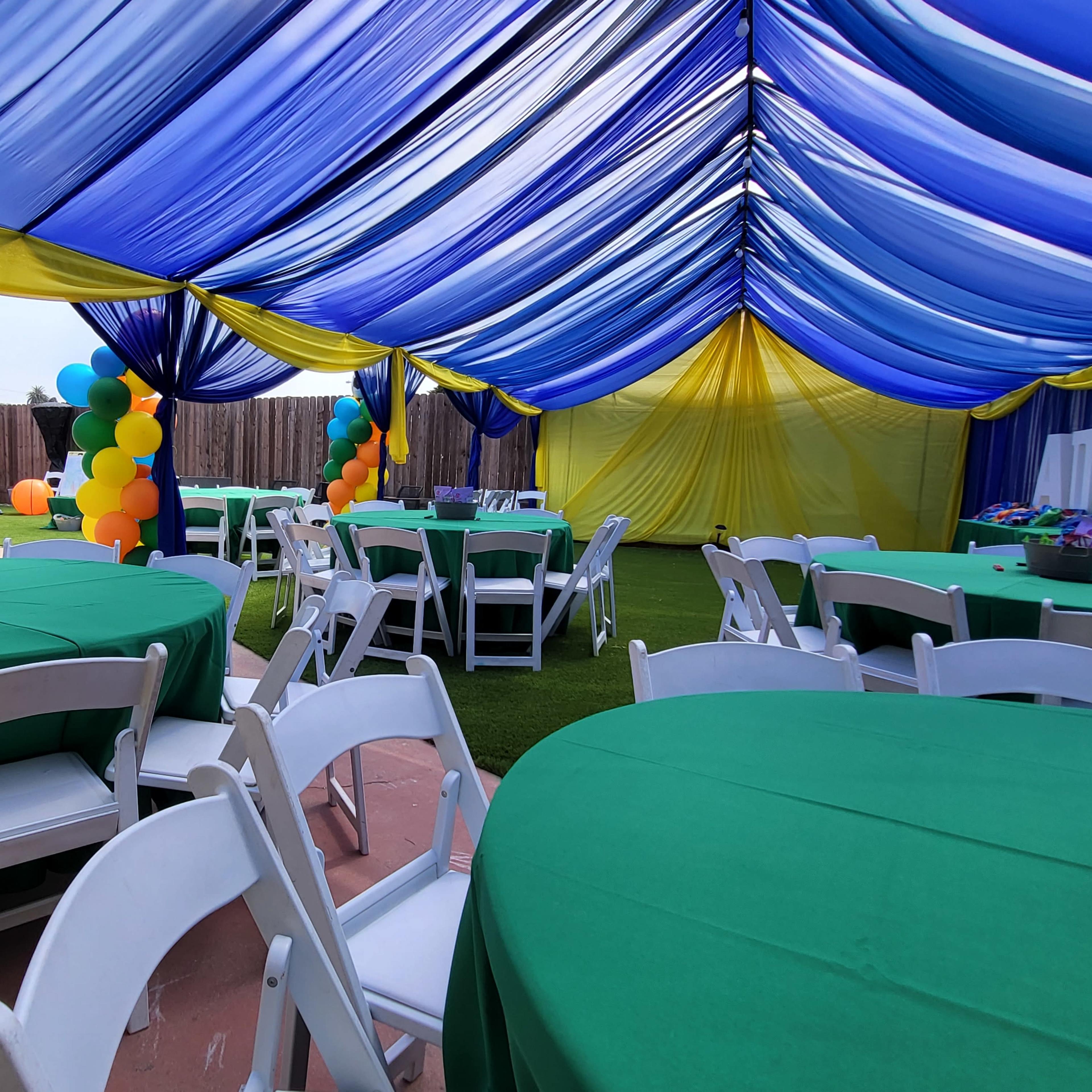 A festive outdoor party setup featuring green tables under a blue and yellow canopy, with colorful balloon decorations and a children's play area in the background.