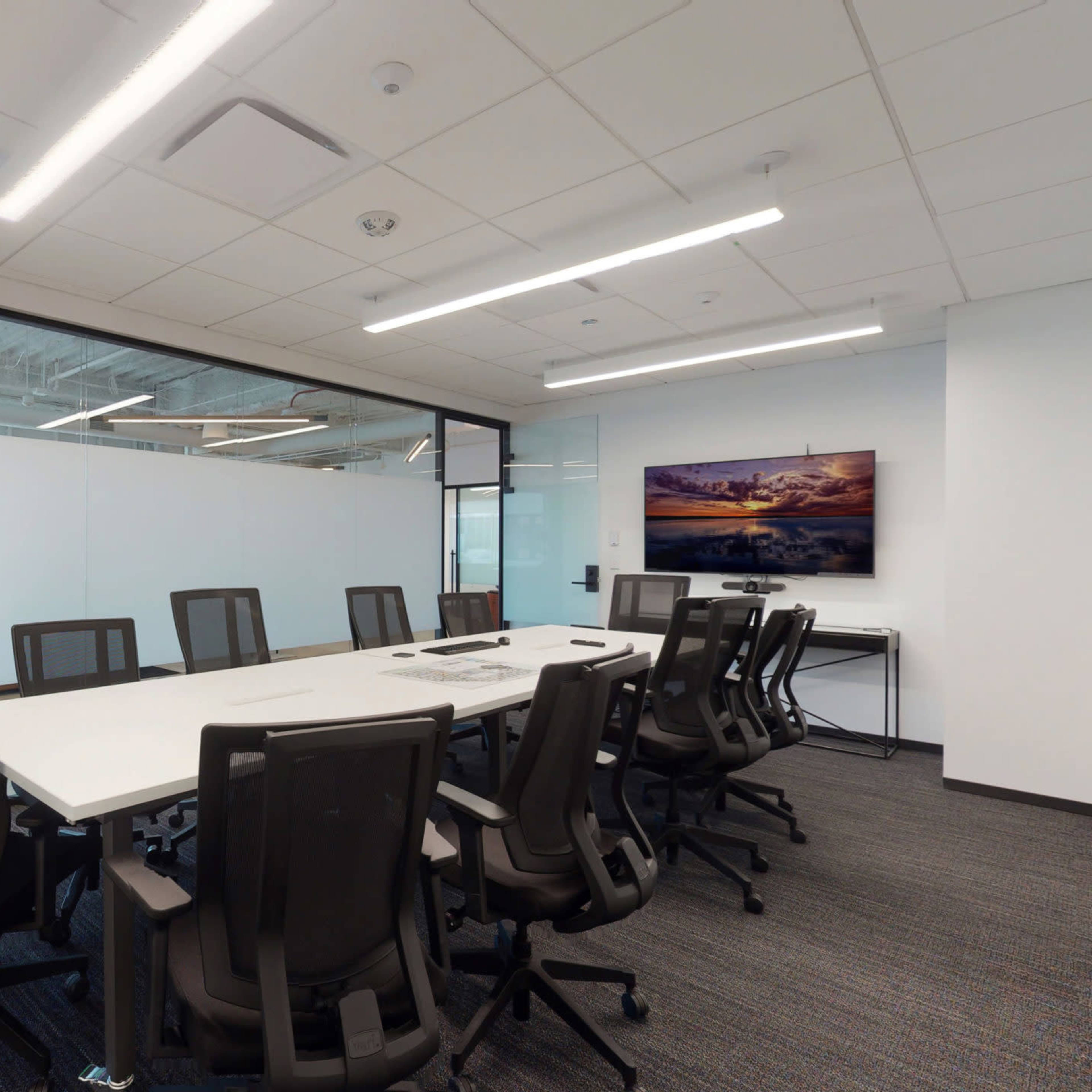 A modern conference room with a long white table surrounded by black chairs, glass walls, and a mounted artwork on one side.